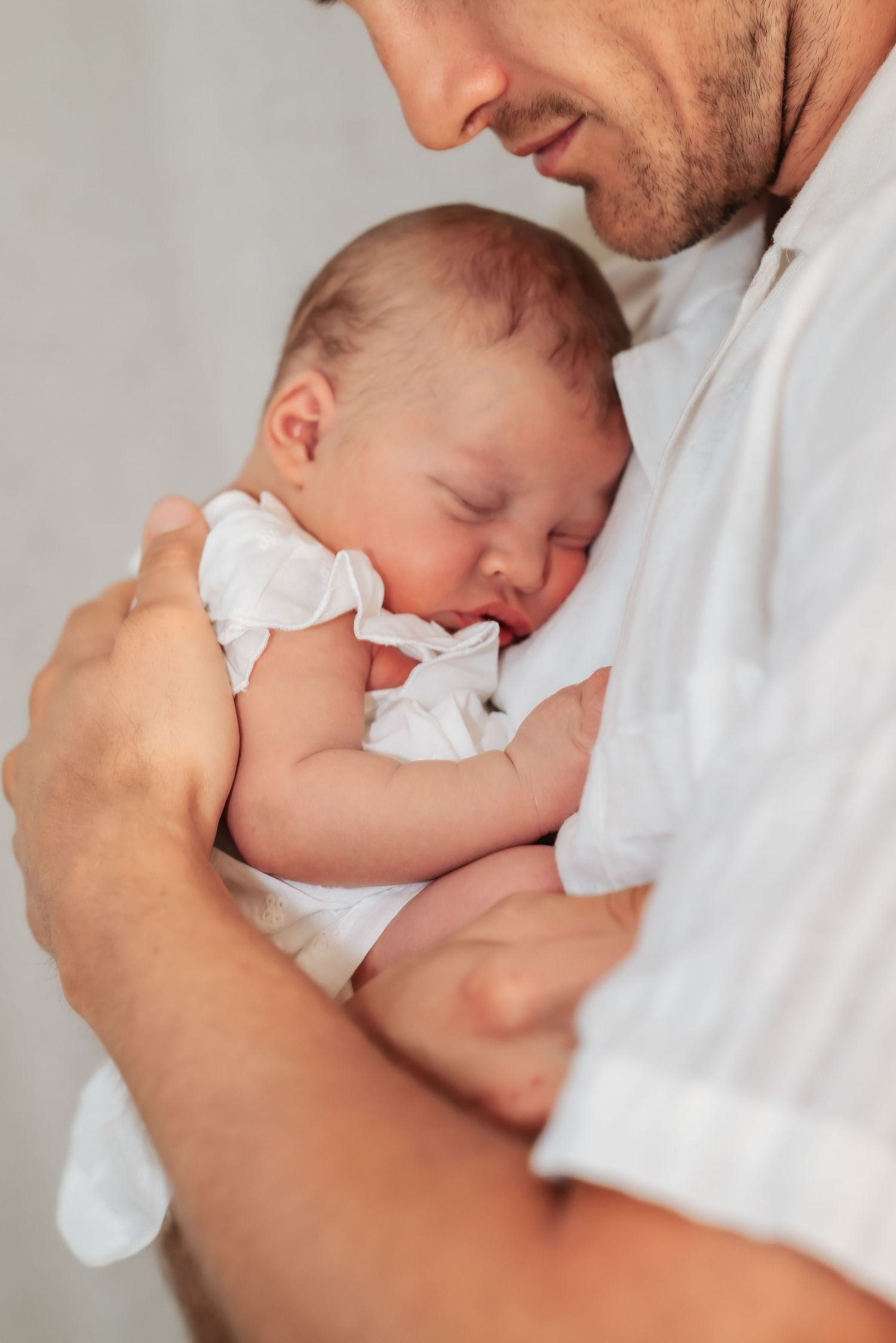 Father gently cradling sleeping newborn baby dressed in white against his chest.