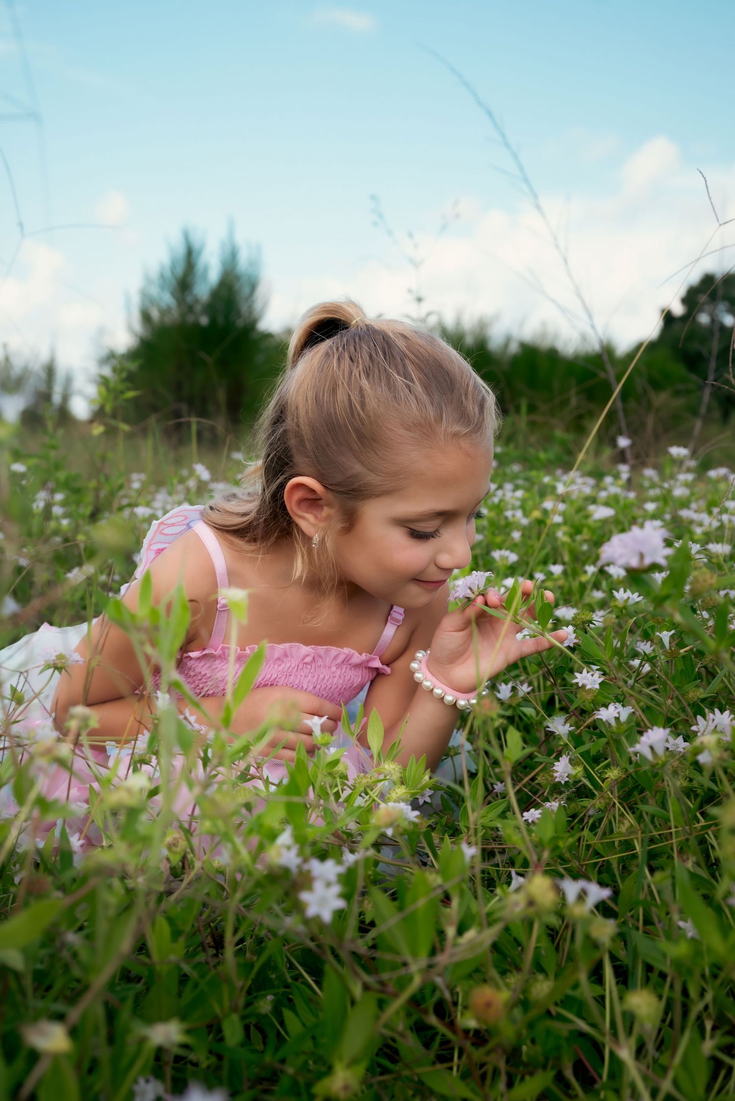 Young girl in pink dress lying in wildflower meadow, smelling small white flowers on a sunny day.