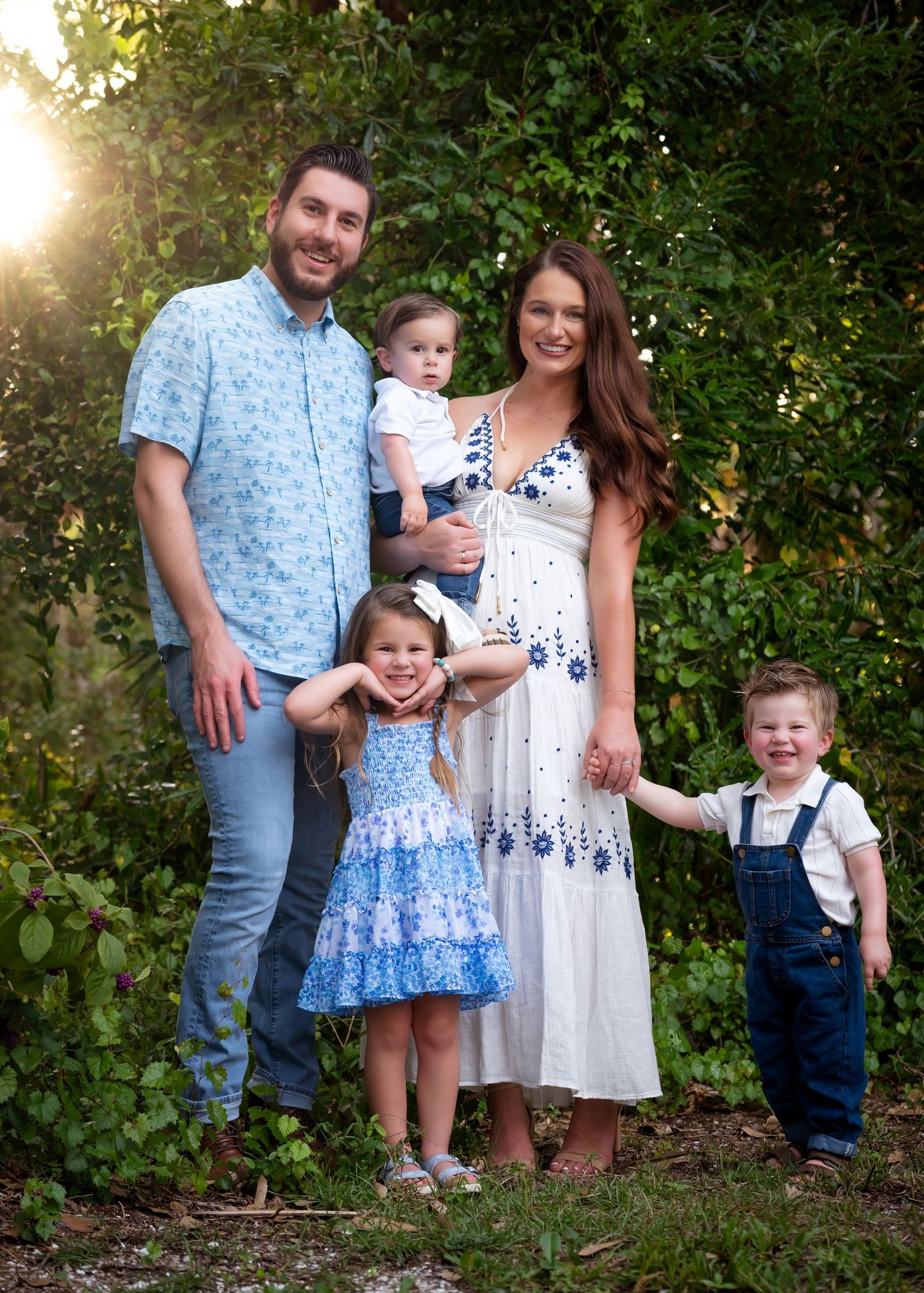 Family of five posing outdoors in coordinating blue and white outfits, smiling together in a lush green garden setting.
