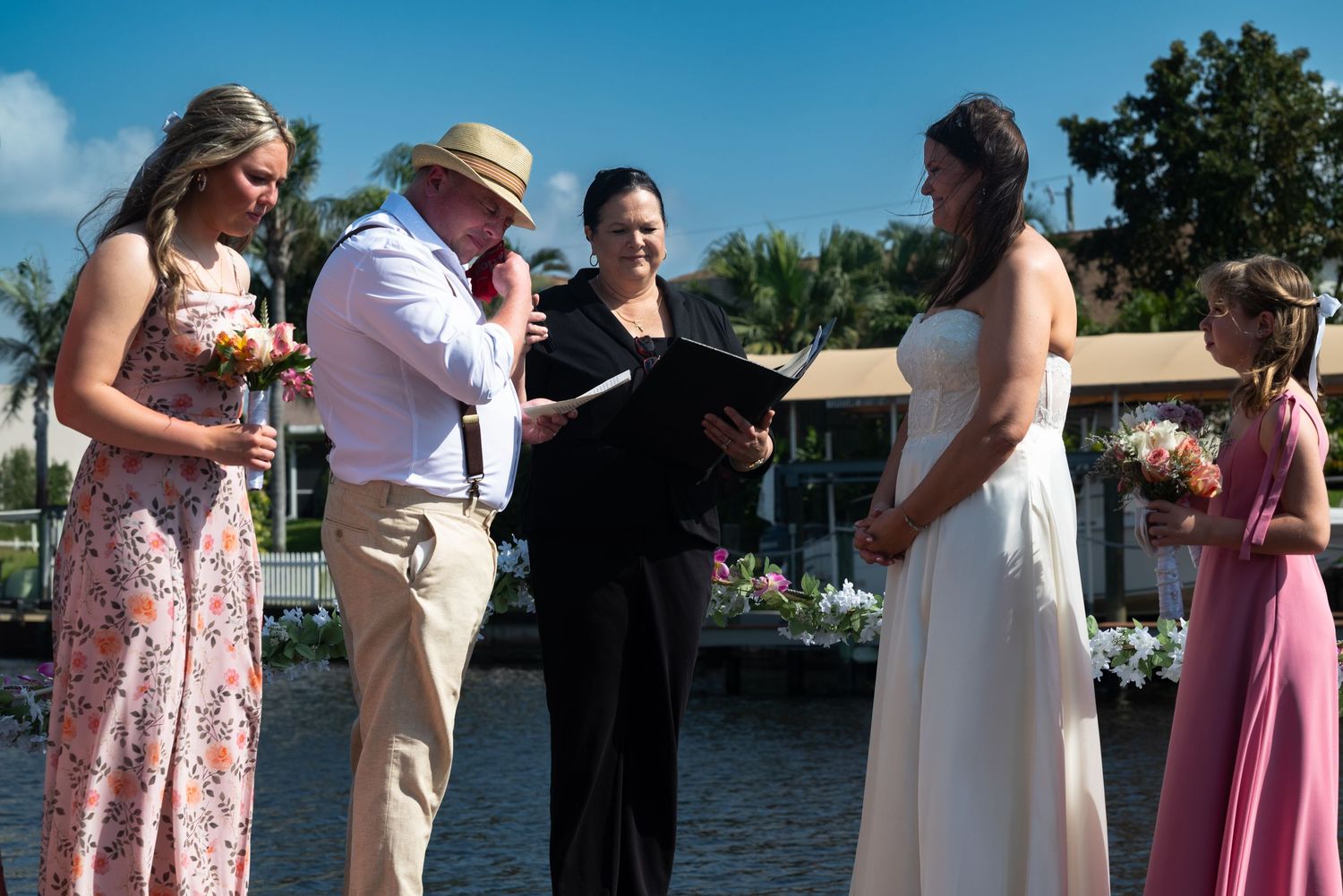 Bride and groom exchange vows with officiant and wedding party during outdoor waterfront ceremony.