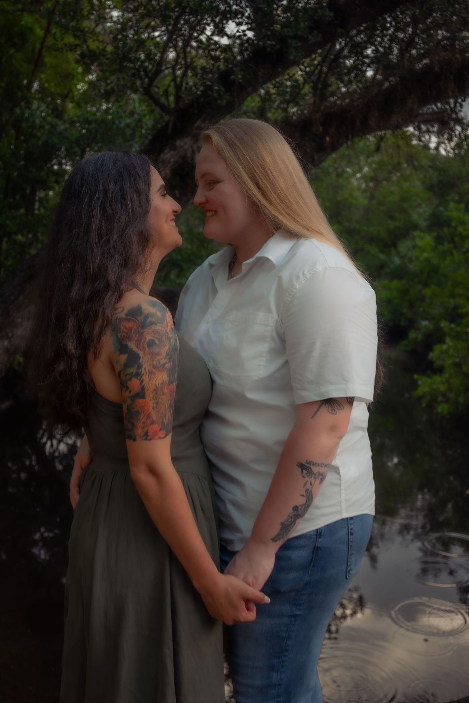 Two women holding hands and smiling at each other near a peaceful riverside setting.
