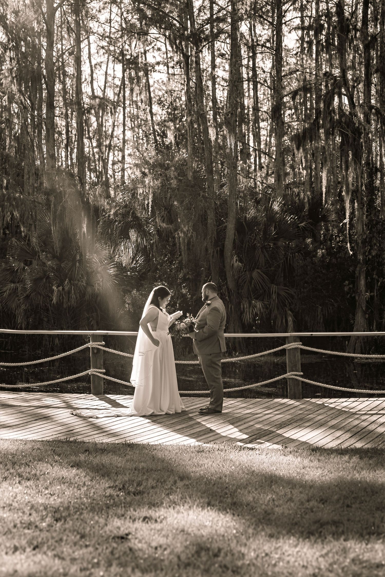 Sepia-toned wedding portrait of bride and groom holding hands on a sunlit wooden deck amid lush forest trees.