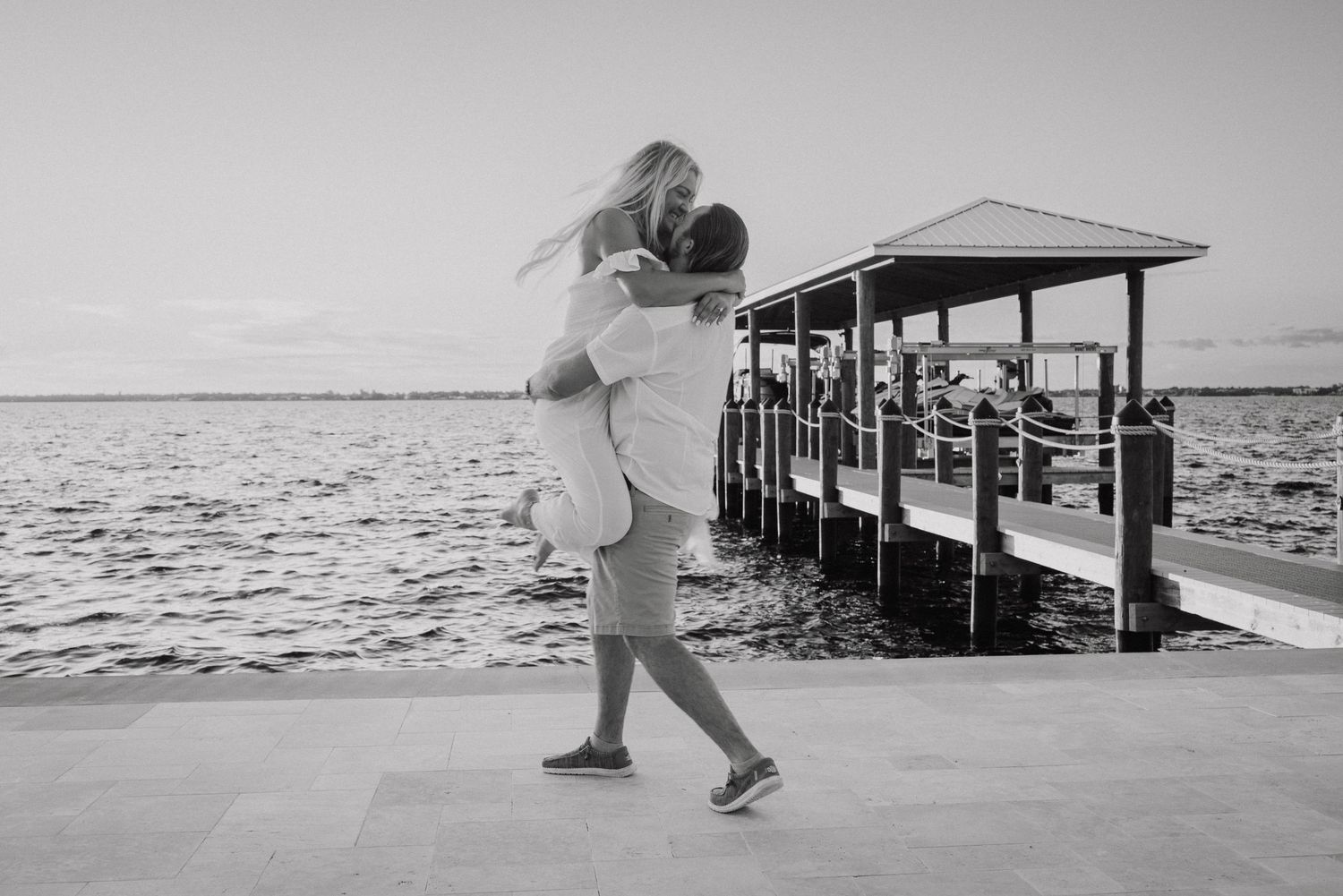 Black and white photo of a couple embracing near a waterfront dock at sunset.