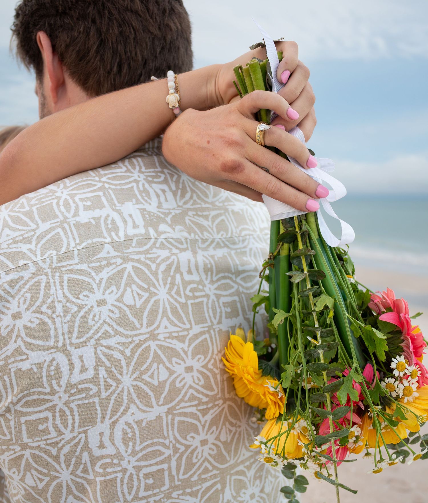 Bride holding colorful wildflower bouquet while embracing groom on beach in patterned shirt.