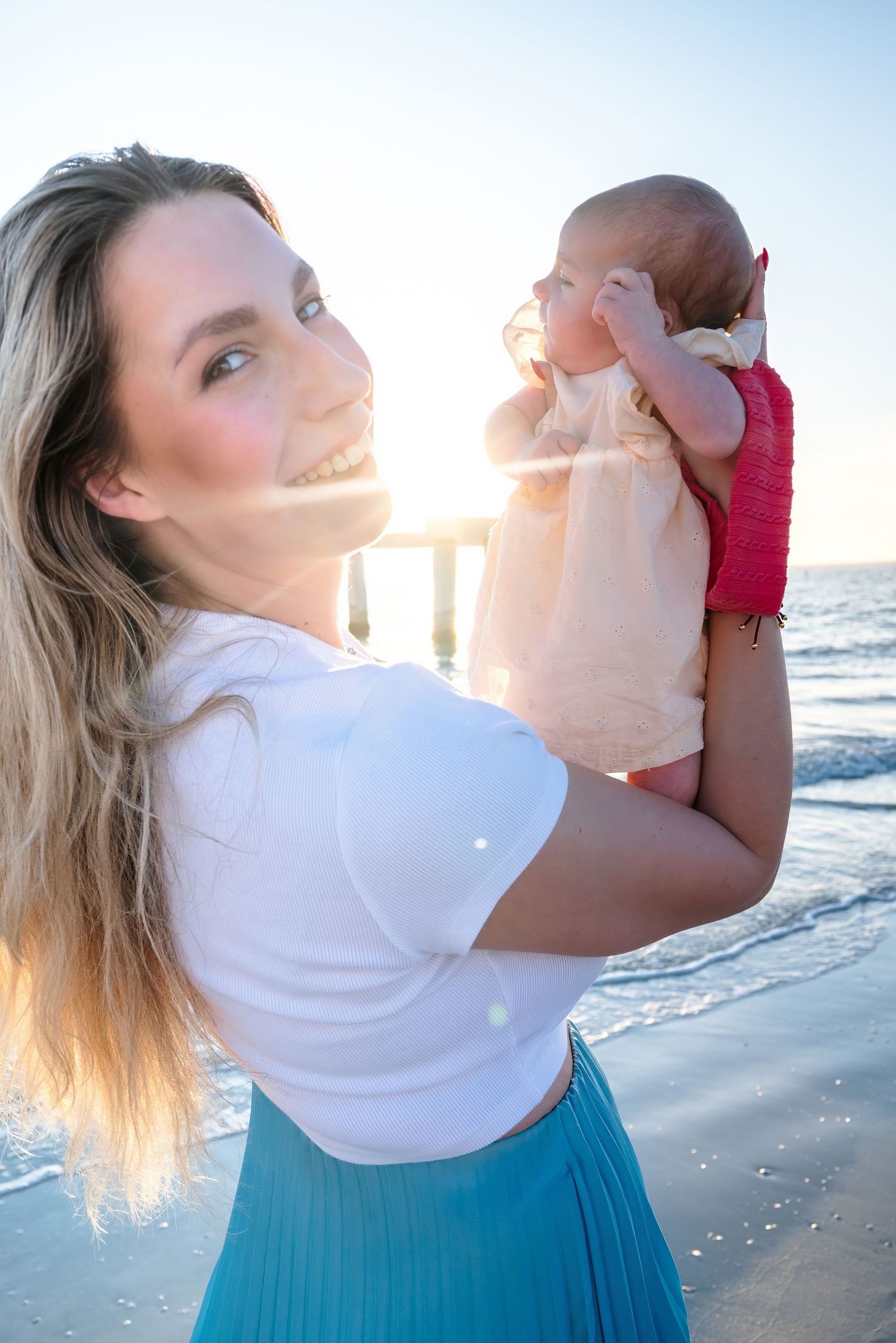 Smiling blonde mother holding newborn baby at golden hour on the beach.