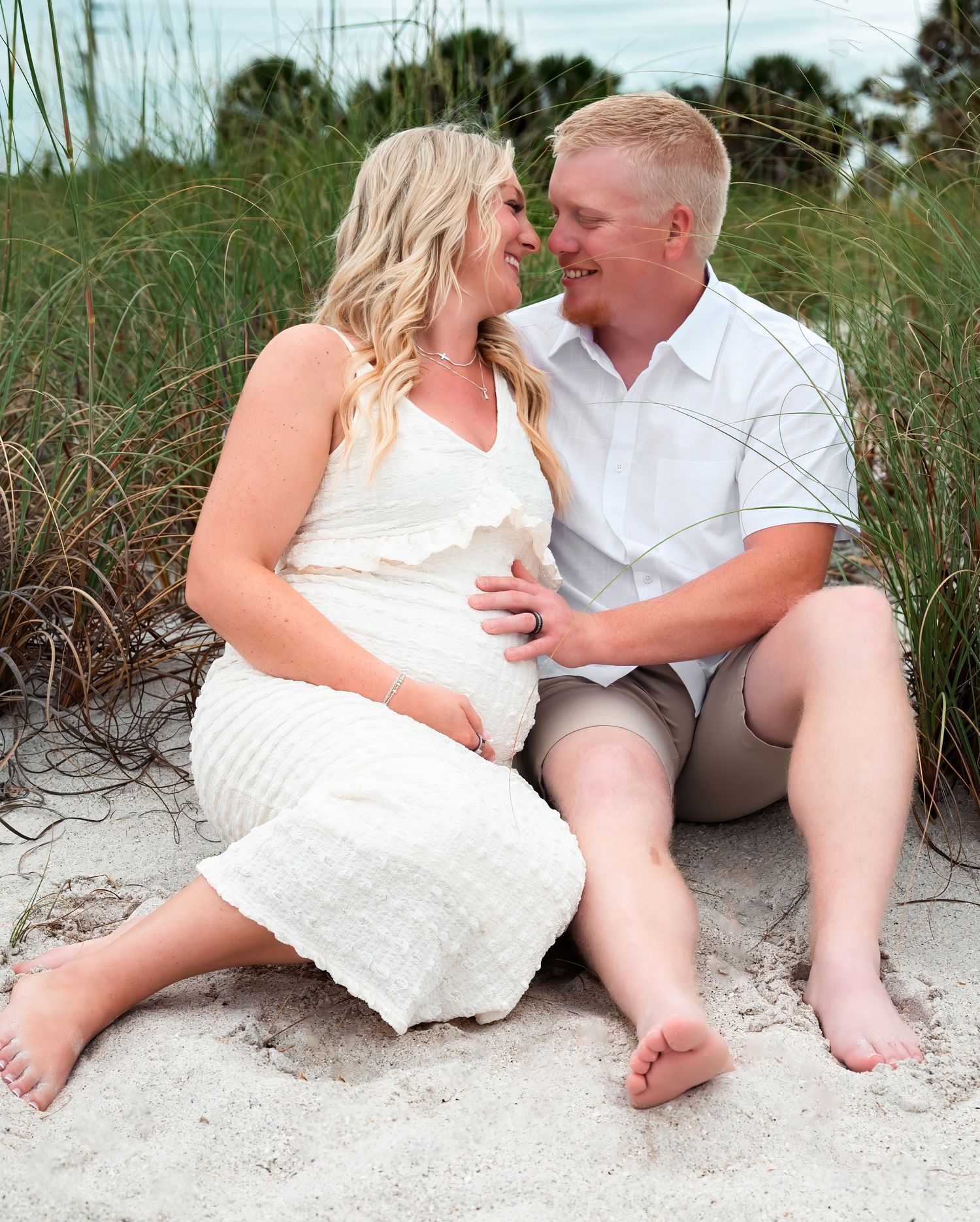 Pregnant couple sitting on sandy beach, woman in white dress cradling baby bump, man in white shirt, smiling at each other.