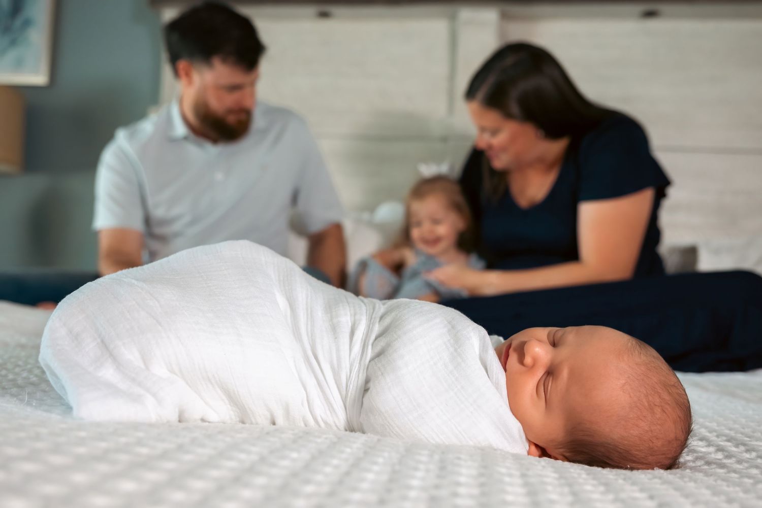 Newborn baby swaddled in white on a bed with parents and toddler sibling smiling in the background.