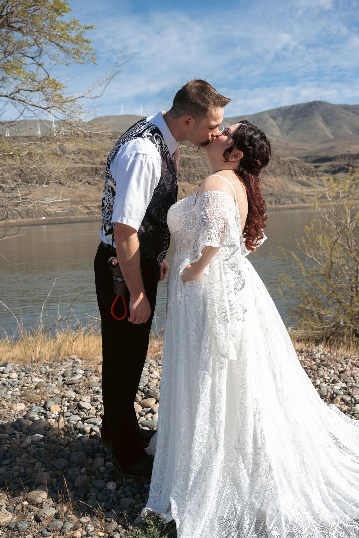 Bride and groom kissing near a rocky lakeside with rolling hills and blue sky in the background.