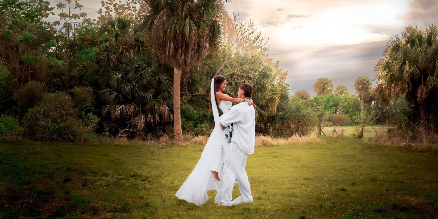 Romantic couple in wedding attire share a joyful embrace on a green lawn with tropical trees at sunset.