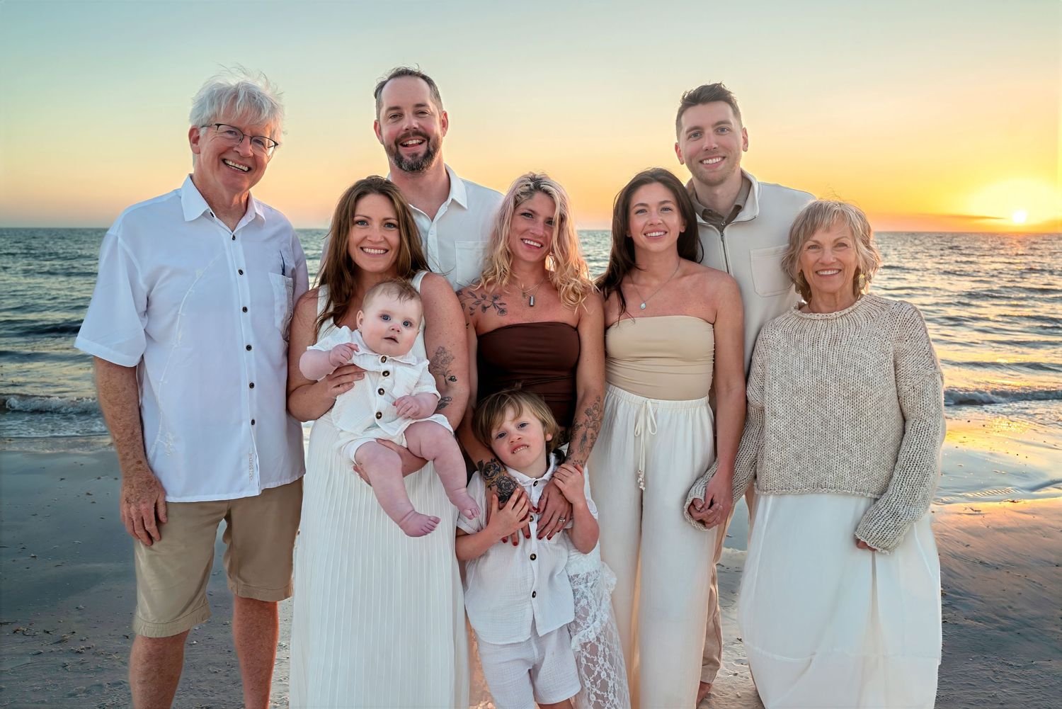 Large multigenerational family group portrait on beach at vibrant sunset in neutral coordinated outfits.