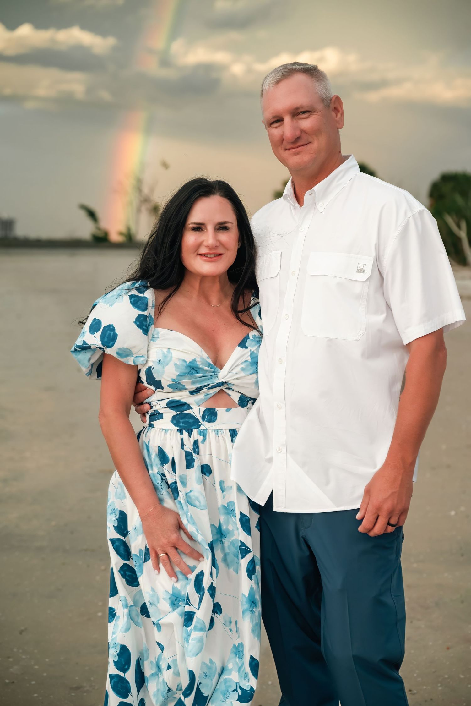 Couple posing on beach with rainbow in cloudy sky, woman in floral dress, man in white shirt and teal pants.