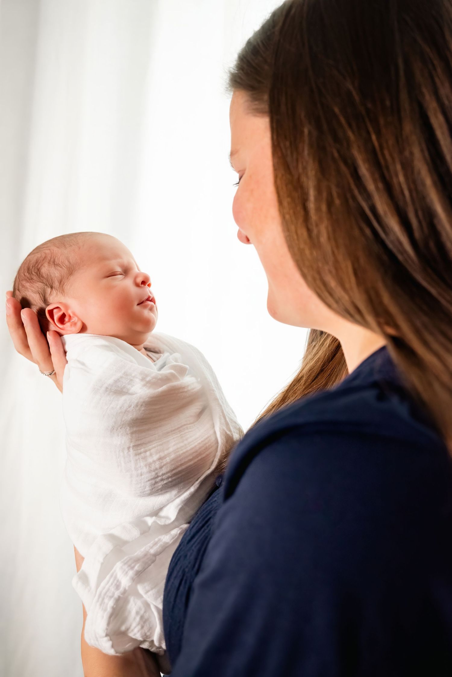 A mother tenderly holds her sleeping newborn baby wrapped in a white swaddle blanket near a bright window.
