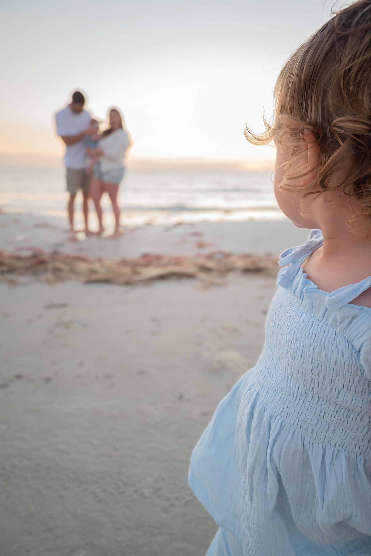 Toddler girl in blue dress in foreground with family embracing at sunset beach in background.