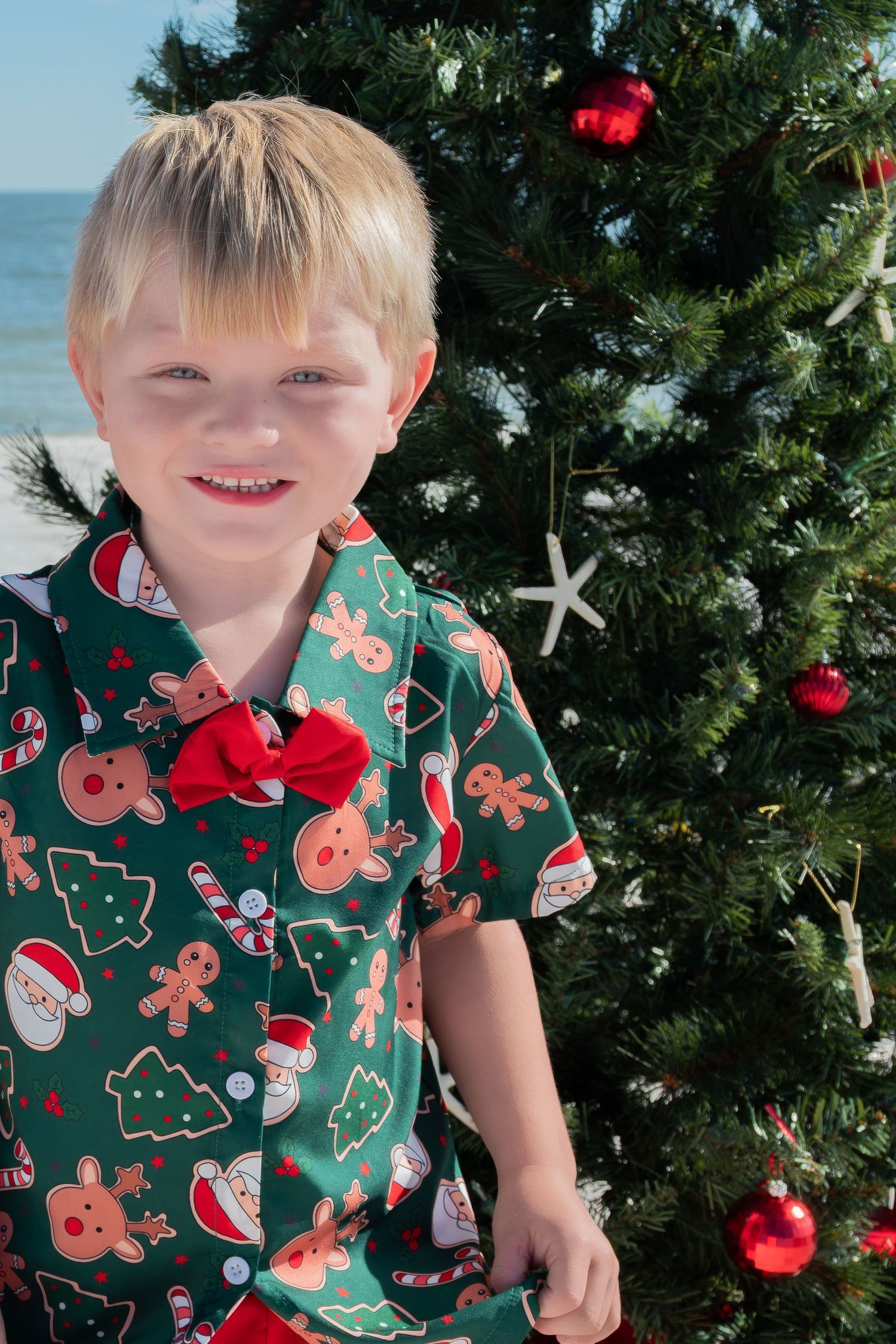 Young boy in green Christmas shirt standing by a decorated beach Christmas tree.