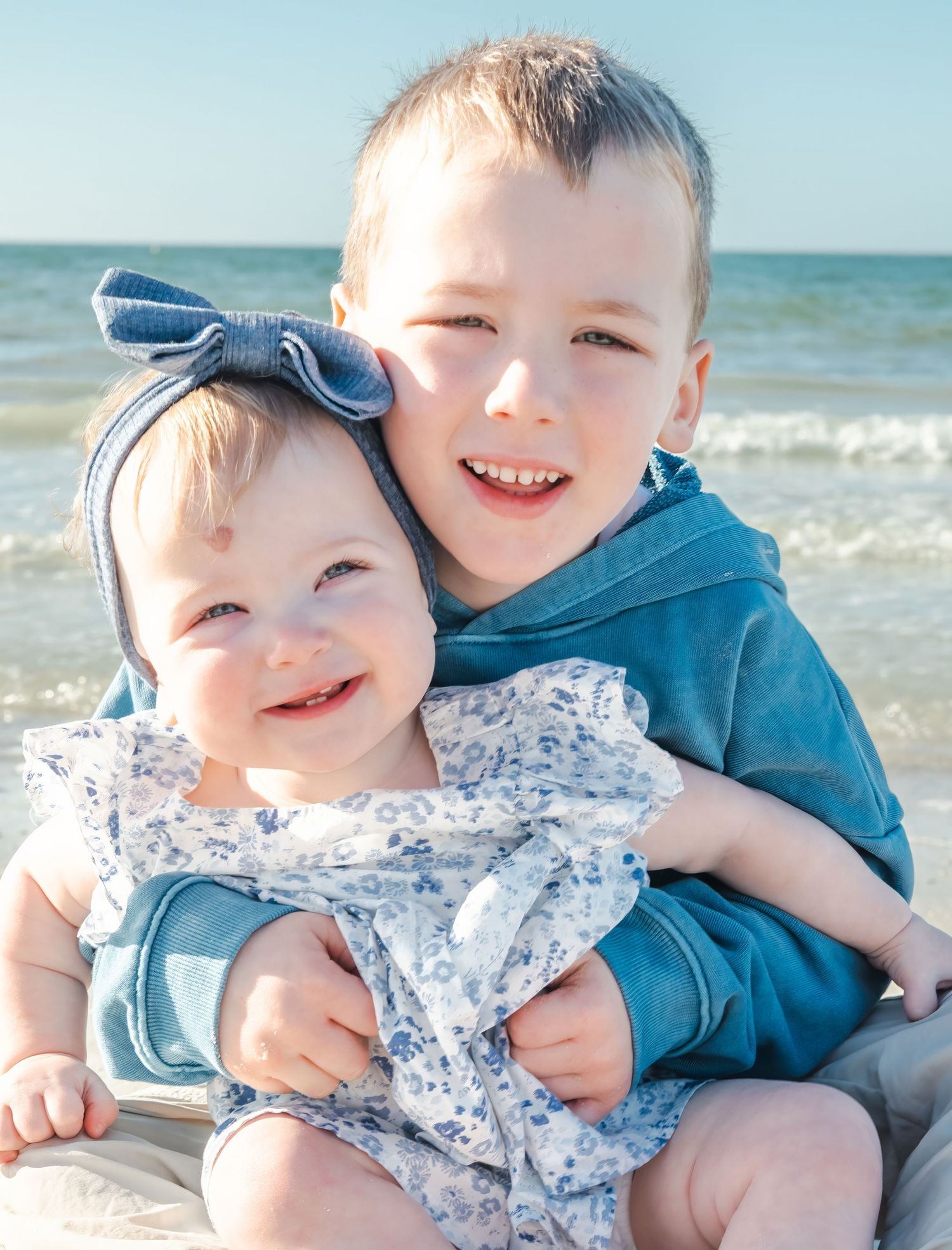 Two young siblings smiling together on a sunny beach, baby girl in floral outfit with bow headband.