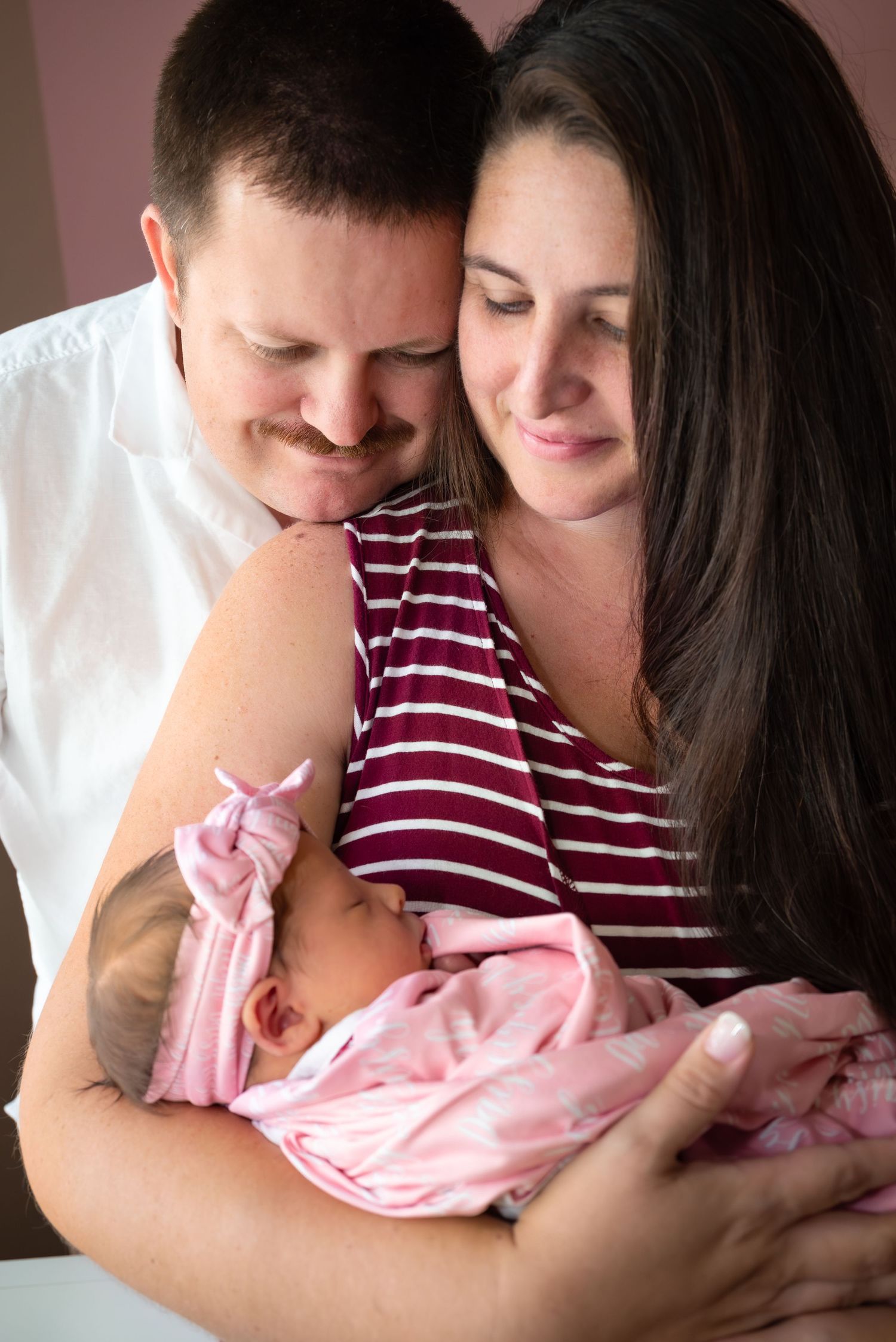 Loving parents smiling down at newborn baby girl in pink swaddle at hospital.