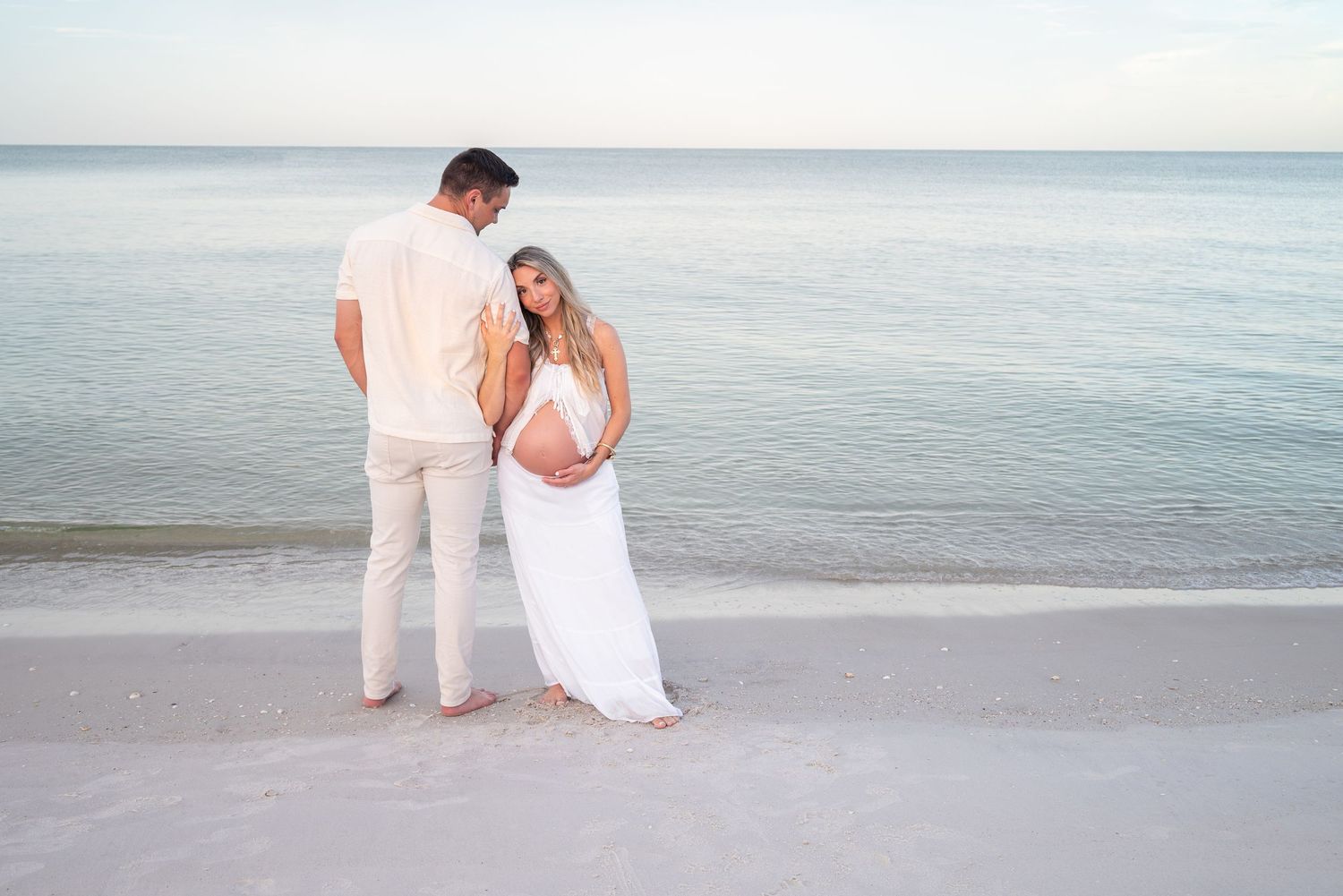 Maternity couple standing at water's edge on a calm beach, woman in white dress holding her baby bump.