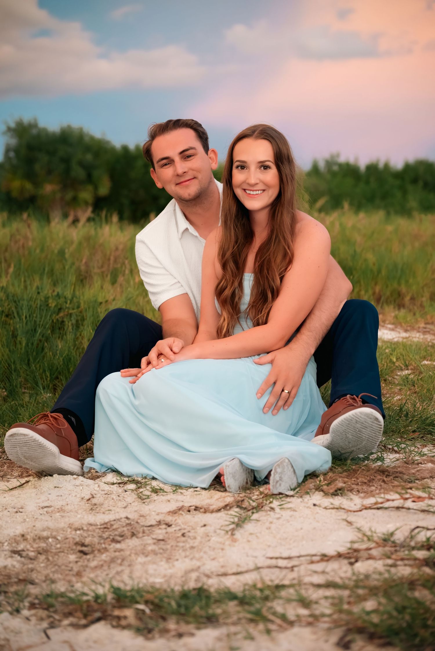 A young couple sits together on sandy ground in a field, smiling, wearing casual attire including a light blue dress.