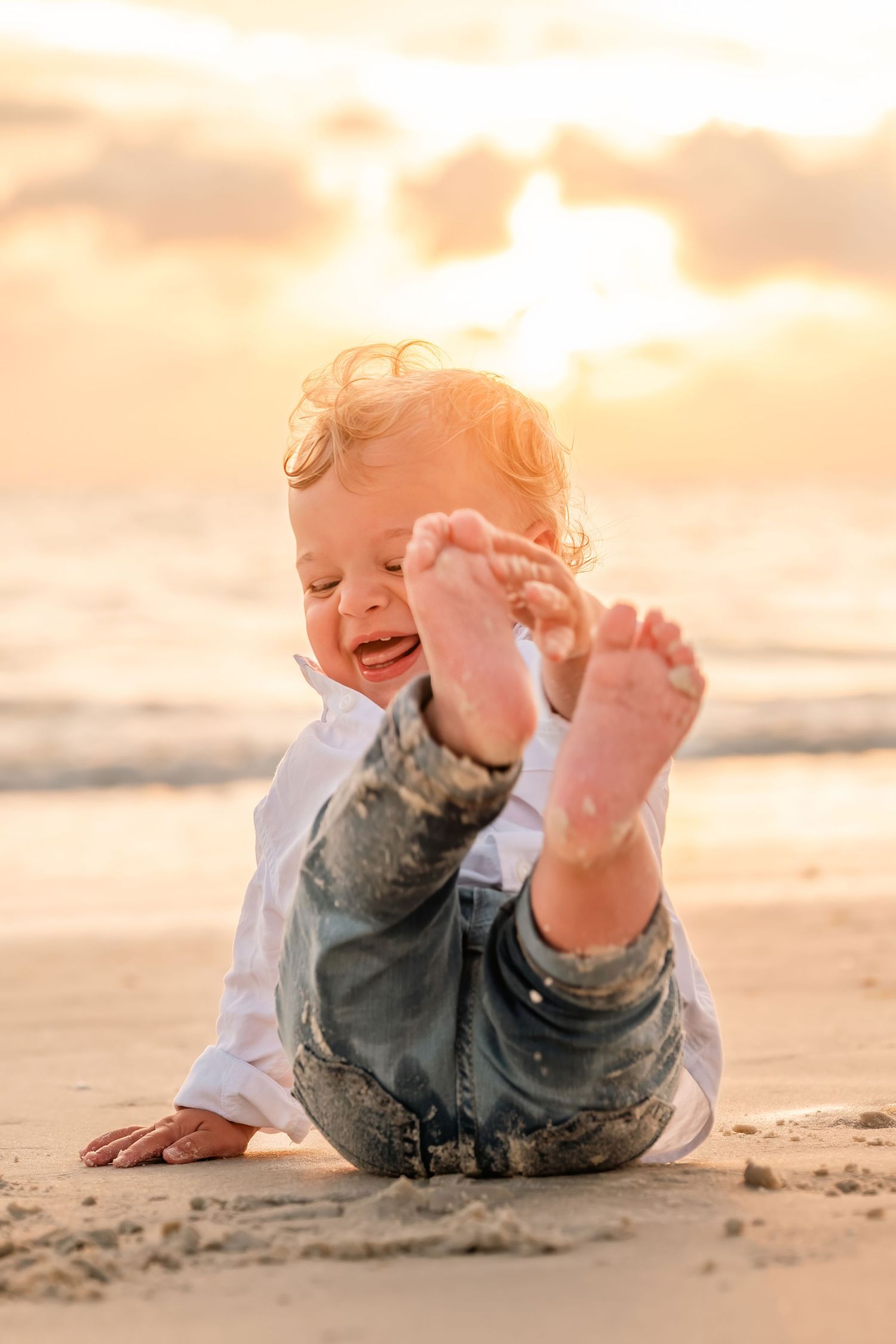 Happy toddler playing with sand on a beach at golden sunset, wearing jeans and white shirt.