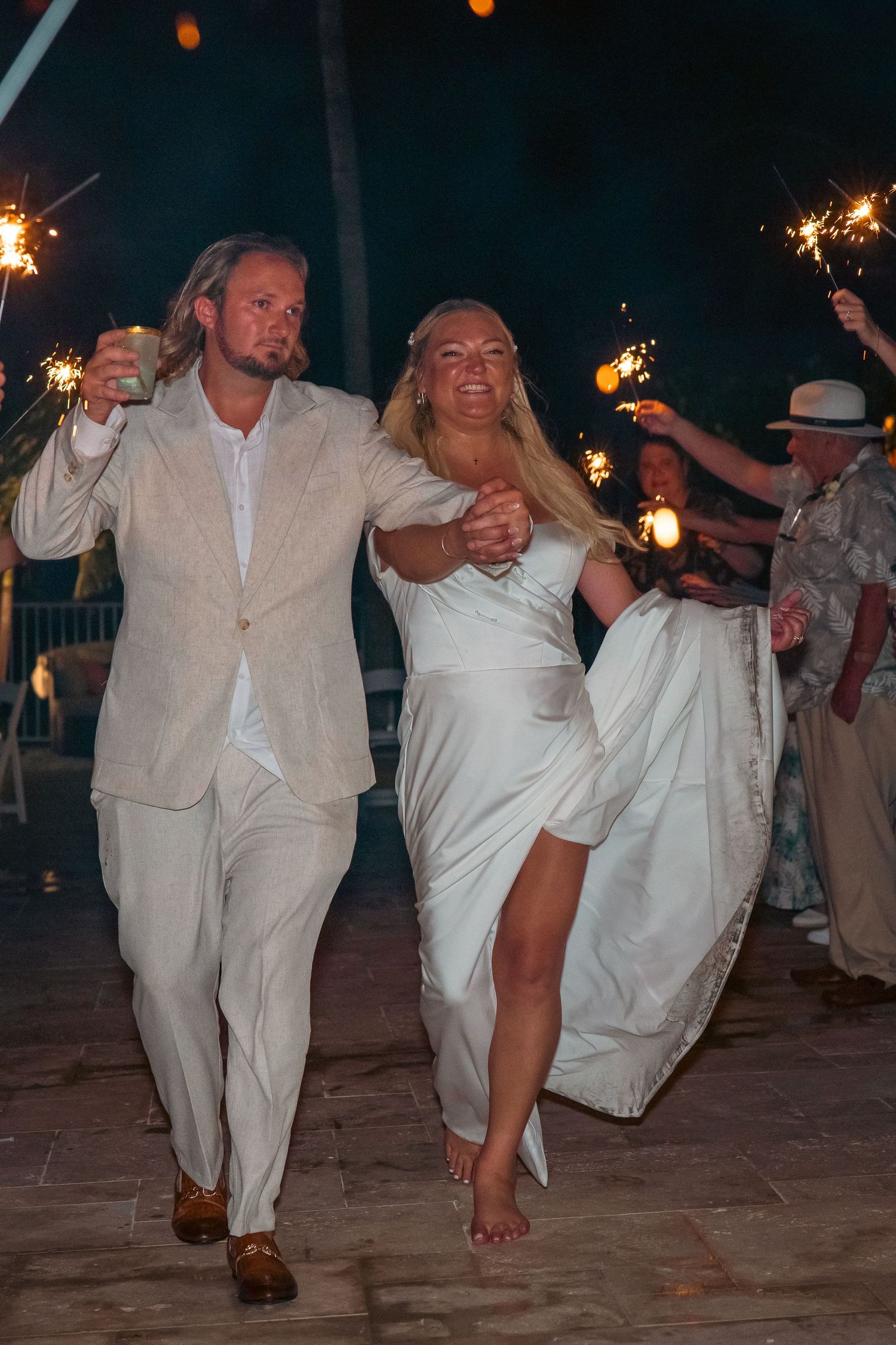 Bride and groom walk through sparkler send-off tunnel during nighttime outdoor wedding celebration.