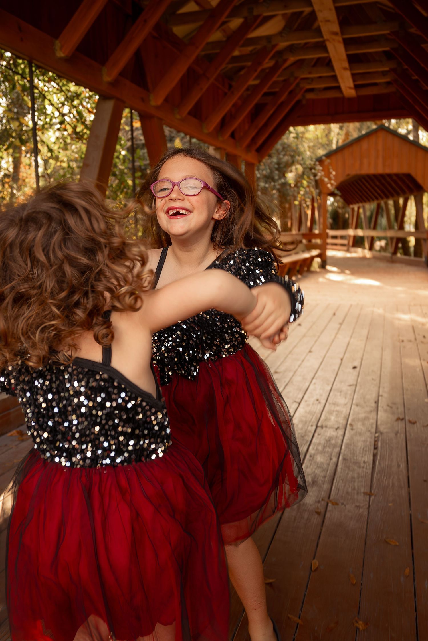 Two young sisters in matching black sequin and red tulle dresses laugh and dance on a wooden bridge.
