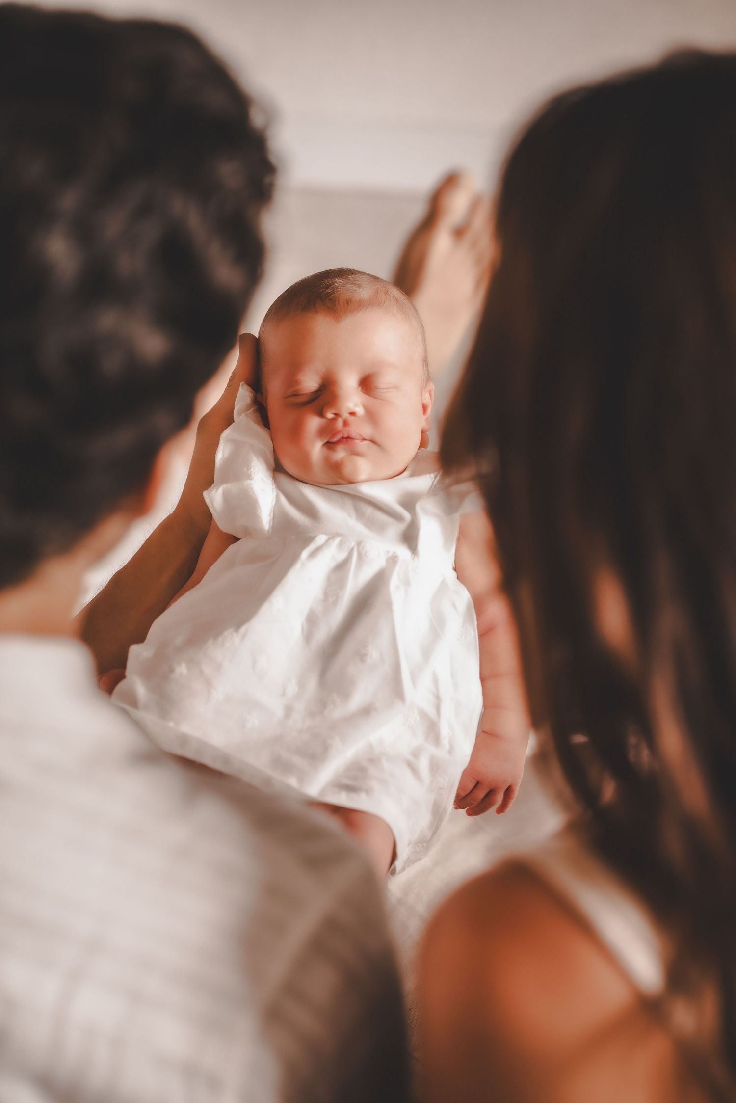 Parents gently hold their sleeping newborn baby in a white outfit between them.
