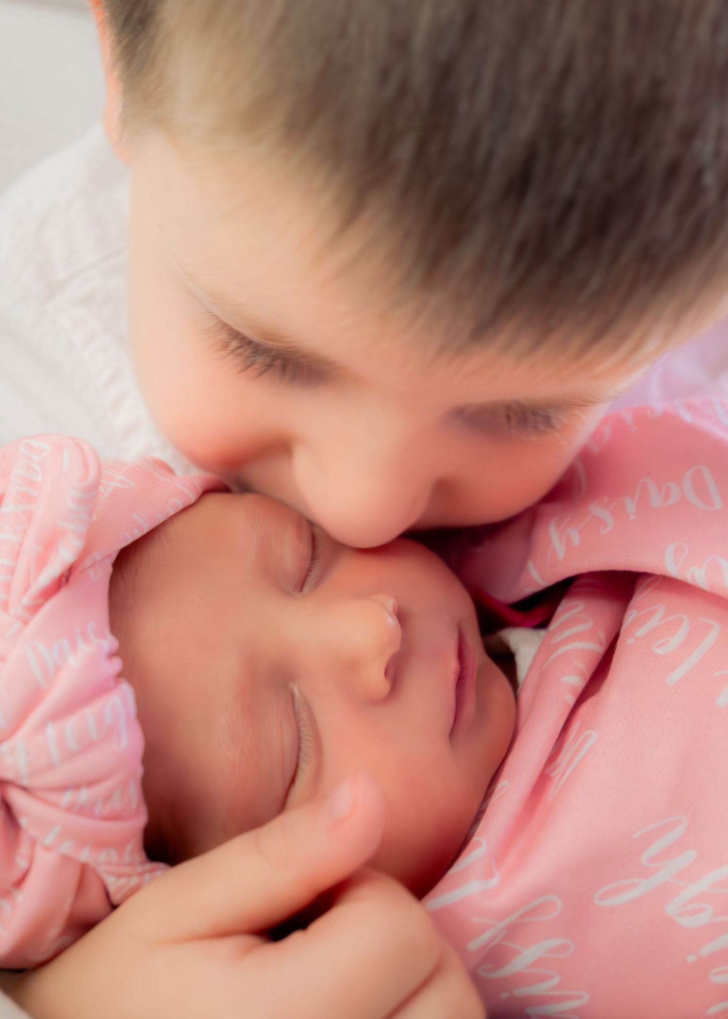Big brother tenderly kisses sleeping newborn baby wrapped in pink swaddle blanket.