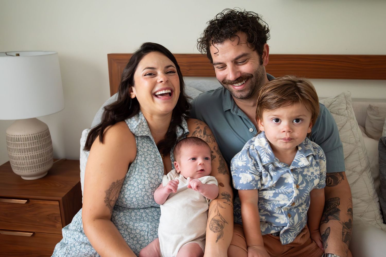 Joyful family of four with newborn baby and toddler posing together on a bed in a bright room.