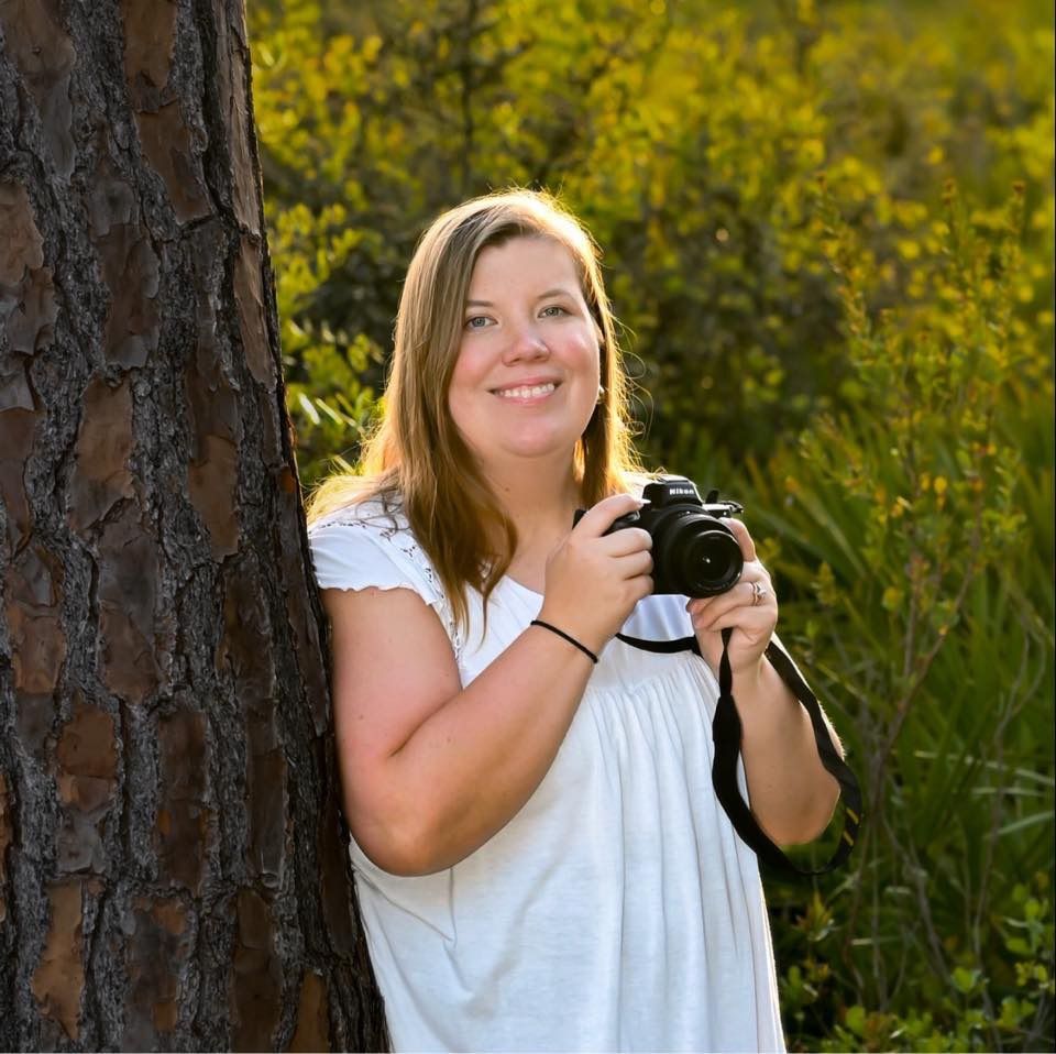 Smiling female photographer holding camera while leaning against pine tree in sunlit outdoor setting.