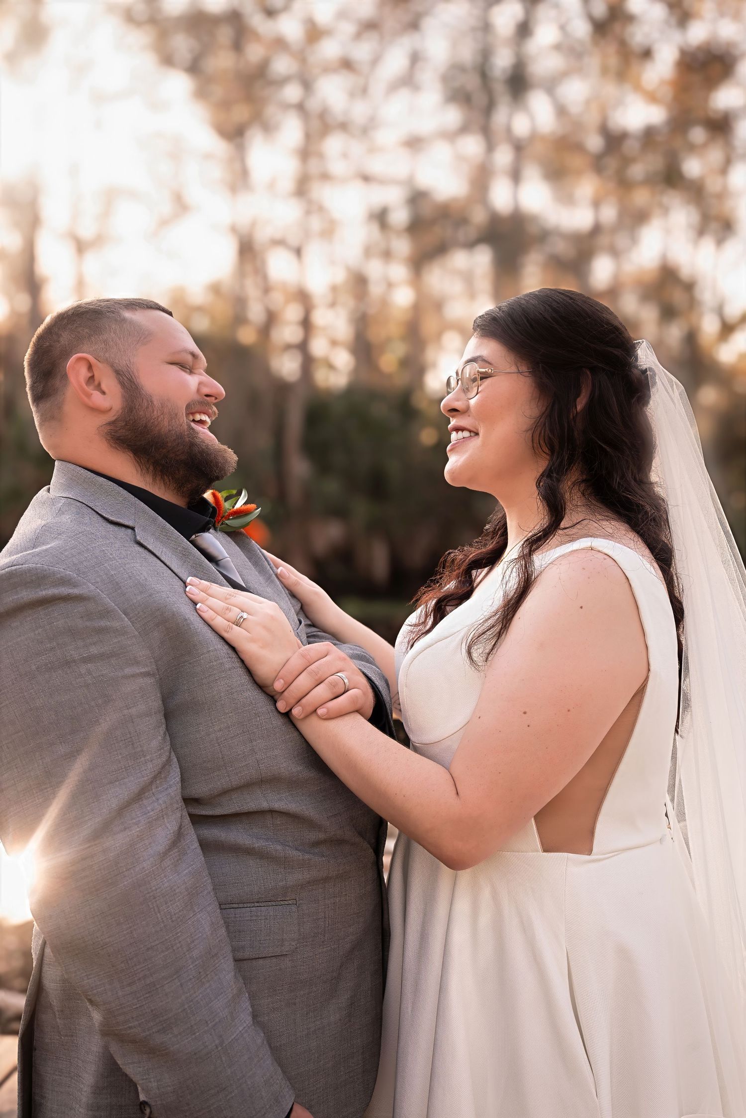 Bride and groom laugh joyfully together outdoors in warm sunset light, bride wearing a sleeveless white gown.