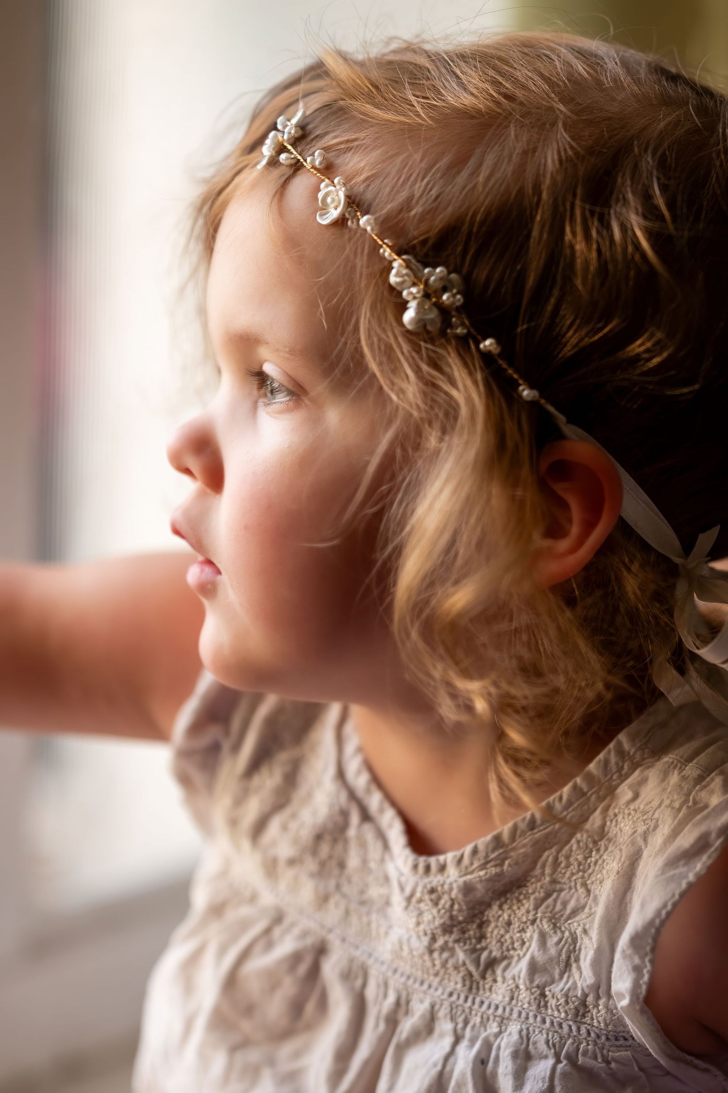 Young girl wearing a delicate pearl headband gazes thoughtfully out a window in soft natural light.