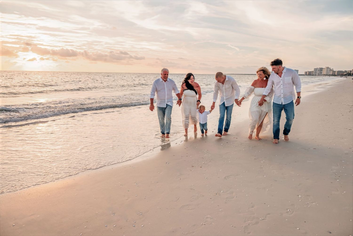 Multi-generational family in white outfits walking hand-in-hand along a sandy beach at sunset.