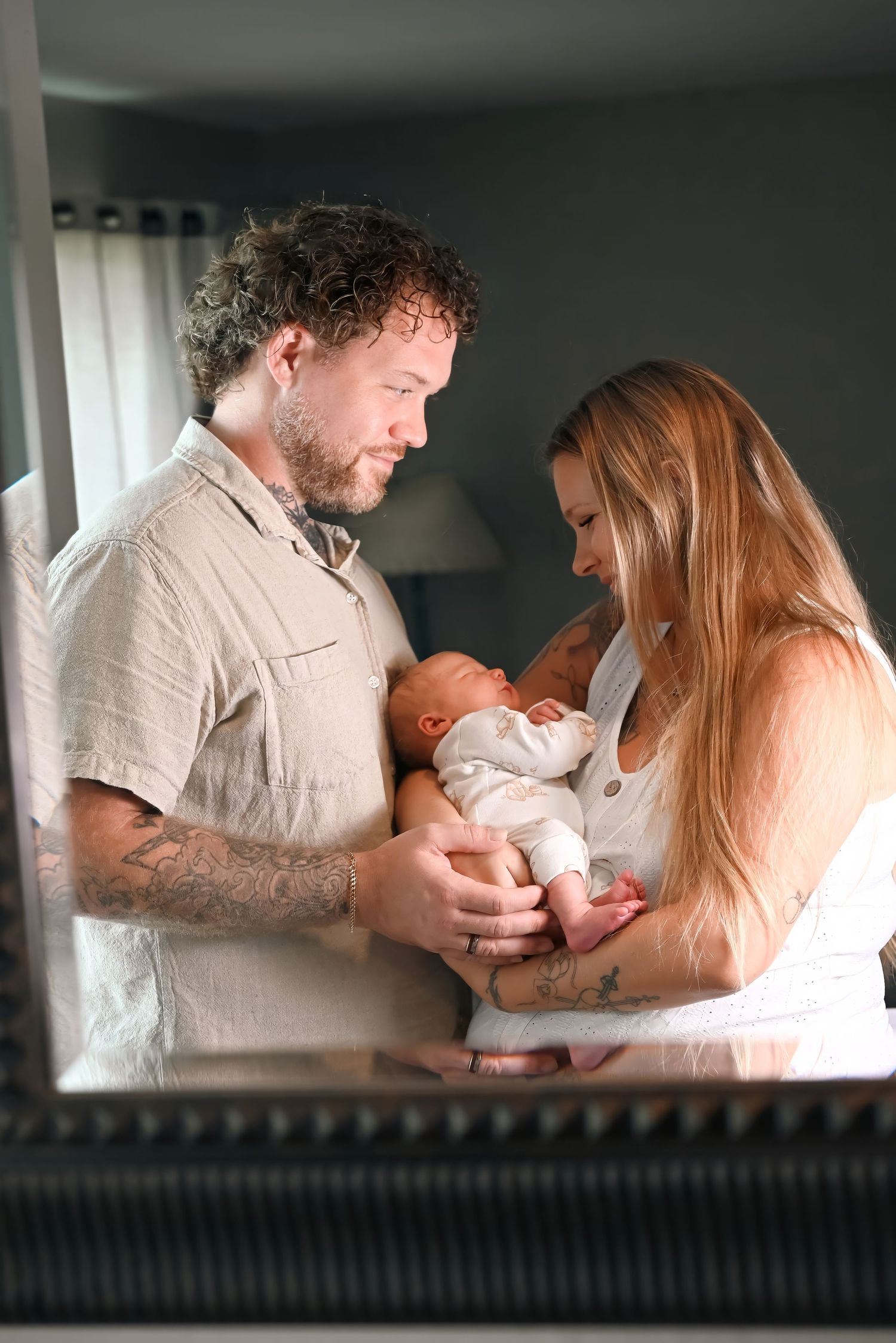 A couple with tattoos tenderly holds their newborn baby together, reflected in a decorative mirror indoors.
