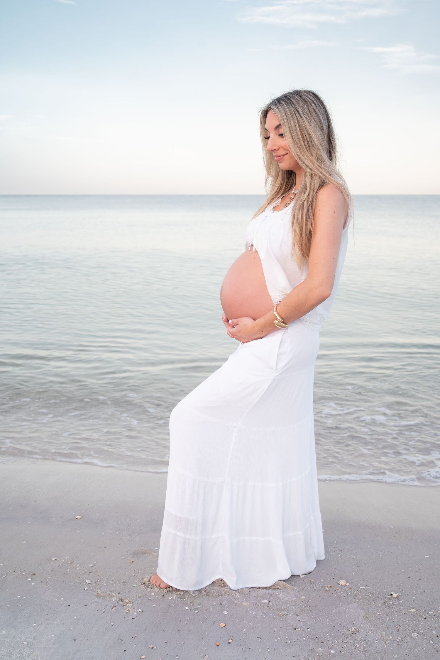 Pregnant woman in white maxi dress standing on beach at sunset, gently holding her bare baby bump.