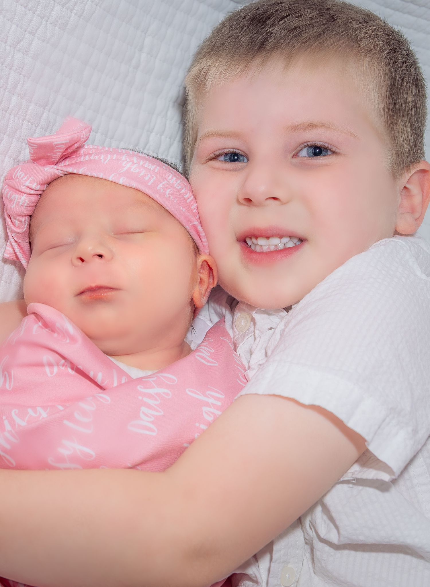 Smiling boy with blue eyes gently holds sleeping newborn sister in pink swaddle.