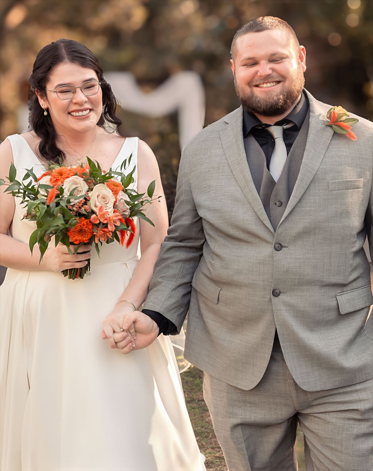 Smiling bride with orange bouquet and groom in gray suit walk after ceremony.