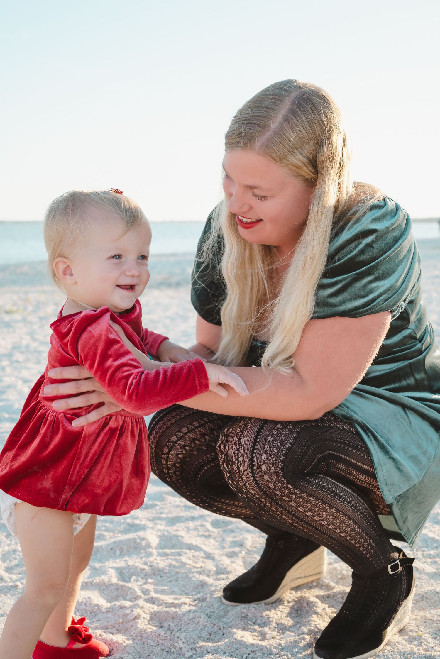 Blonde mother crouching on beach smiling at her happy toddler in a red dress.