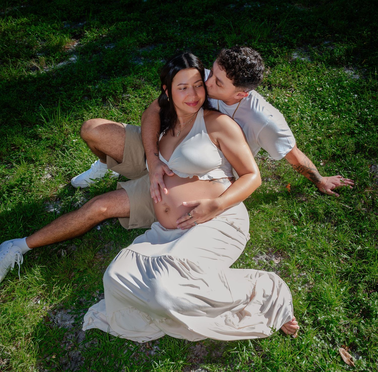 Couple sitting on grass, man kissing pregnant woman's cheek while holding her baby bump, wearing neutral tones.
