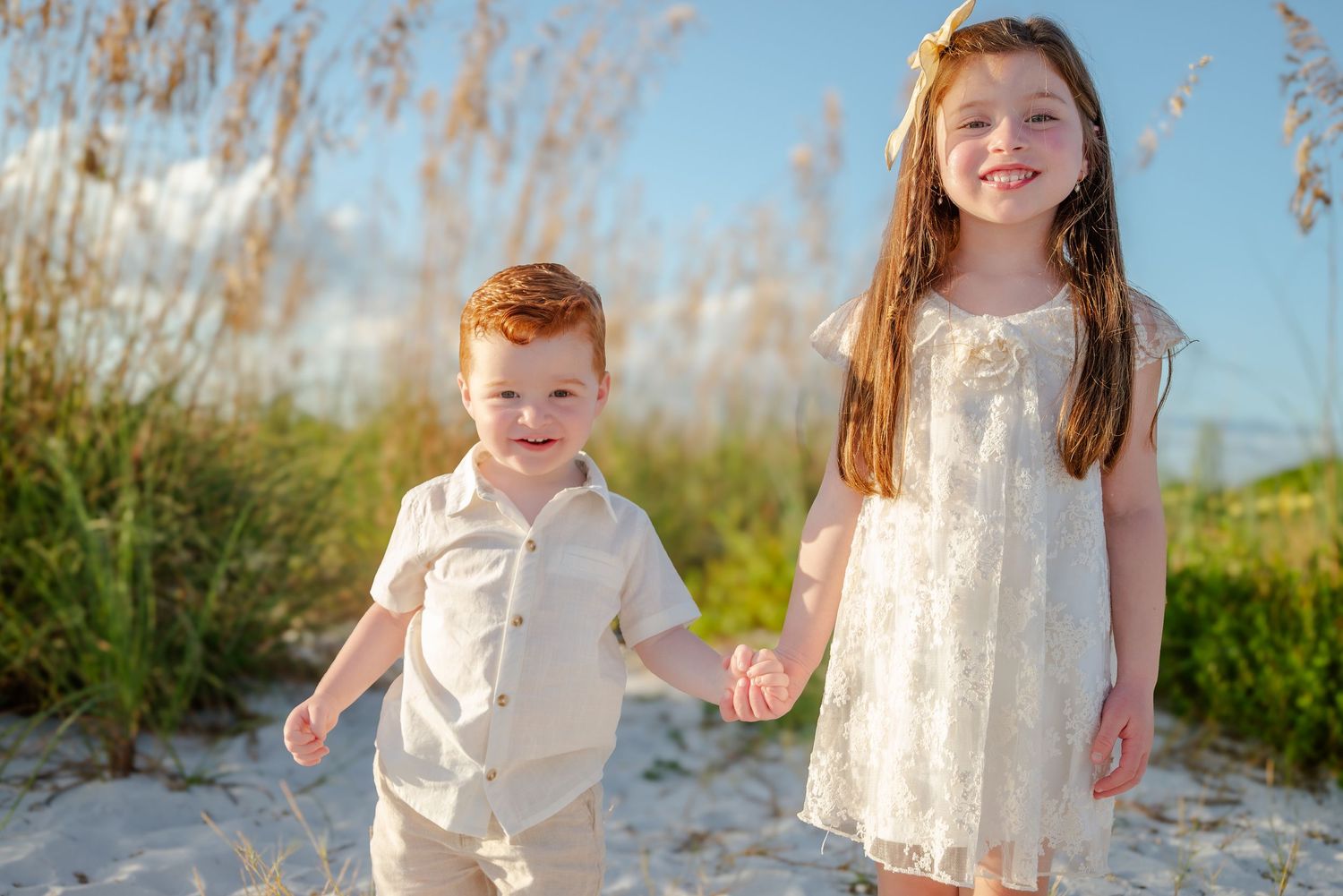 Two young siblings, a redhead boy and brunette girl, holding hands and smiling on a sunny beach.