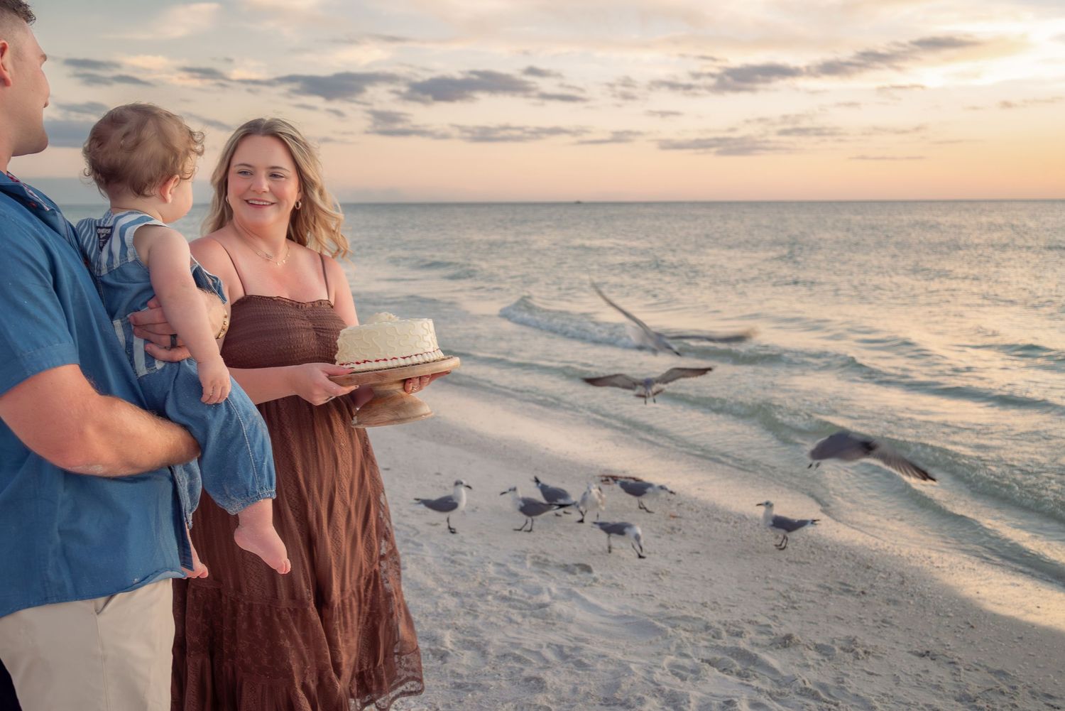 Happy family with baby and birthday cake standing on beach at sunset with seagulls flying nearby.