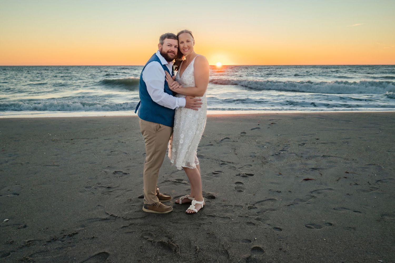 Bride and groom hugging on sandy beach at vibrant orange sunset with ocean backdrop.