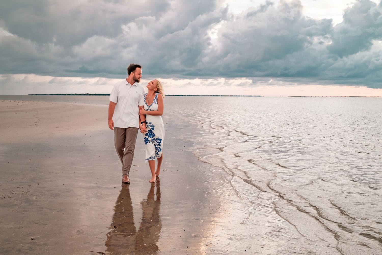 Couple walking hand in hand on a sandy beach with dramatic cloudy skies and ocean waves behind them.