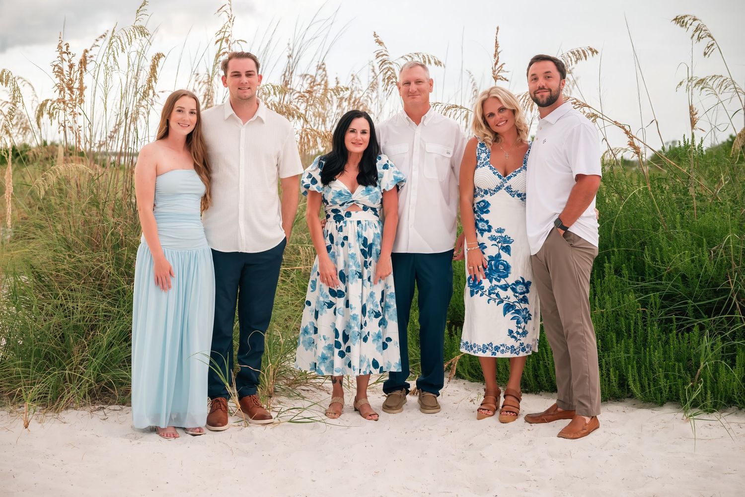 Six people posing together on a sandy beach with sea oats, dressed in elegant summer attire for a family photo.