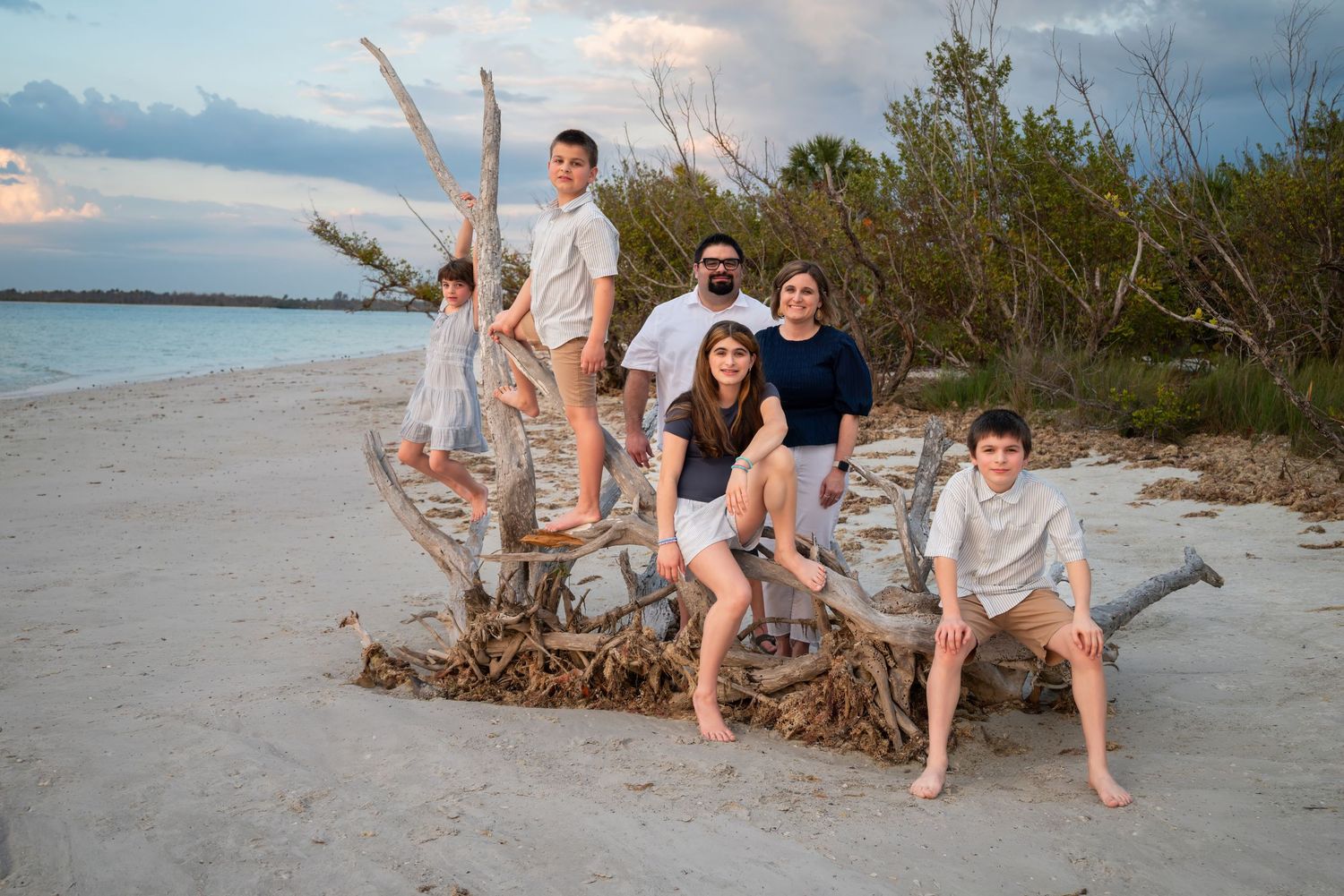 Family of six posing on driftwood at a sandy beach during golden hour with turquoise water and greenery.