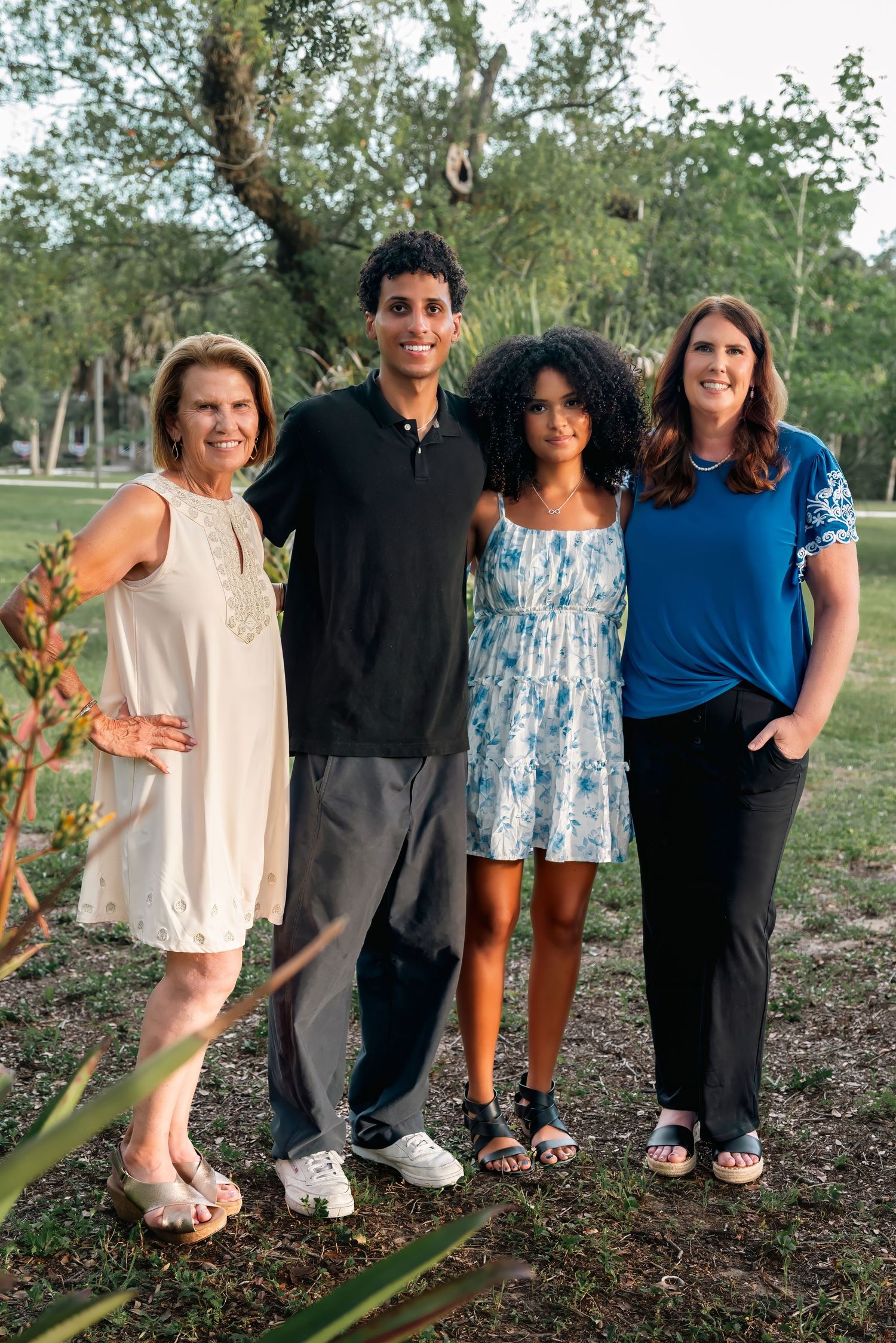 Four people smiling together outdoors in a park setting, dressed in casual attire on a warm day.