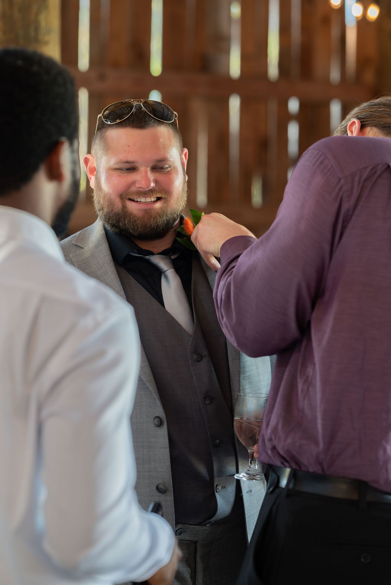 Smiling bearded groomsman in gray suit getting boutonniere pinned inside rustic barn wedding venue.