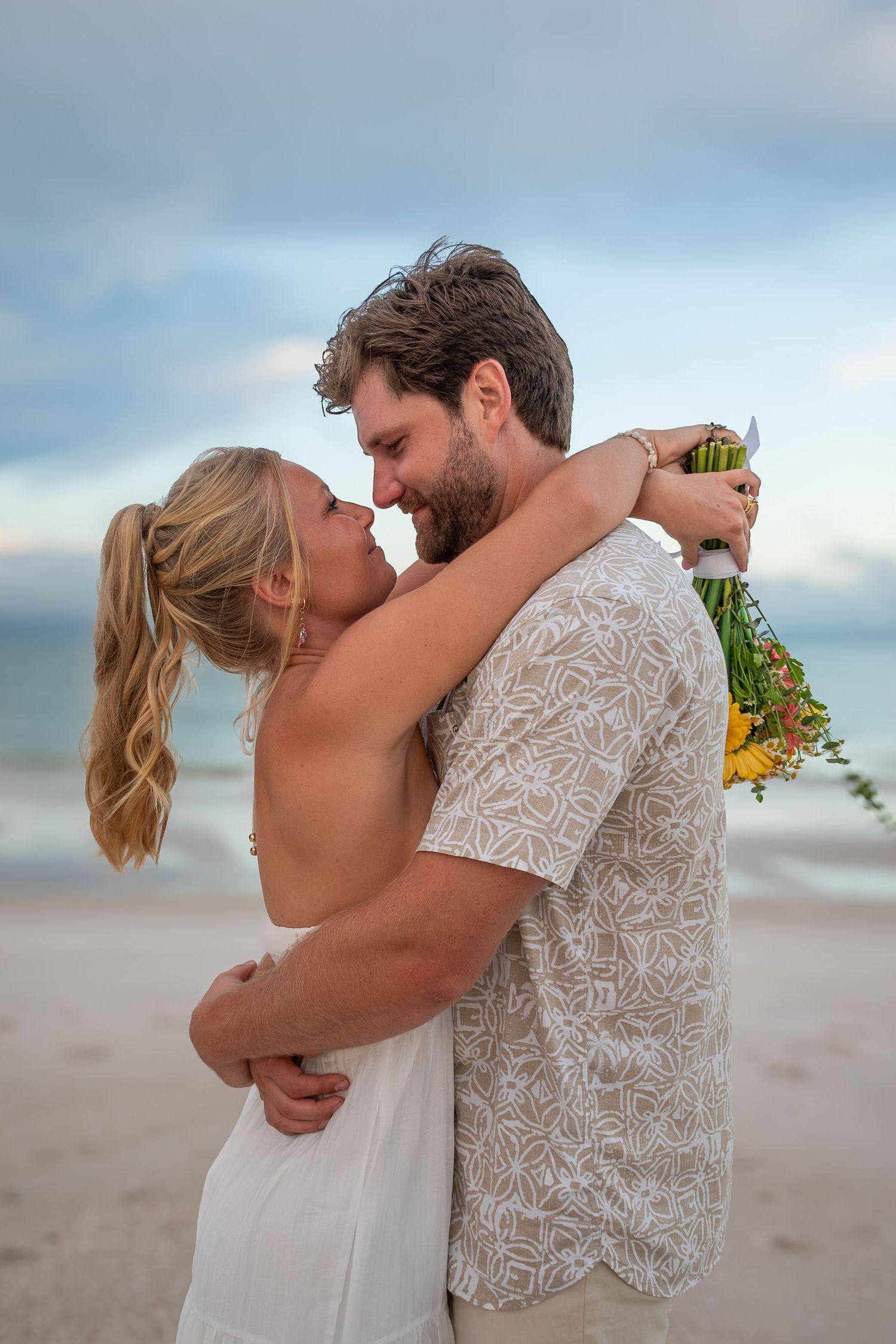 Couple embracing on beach at sunset, woman in white dress holding colorful bouquet, romantic seaside celebration.