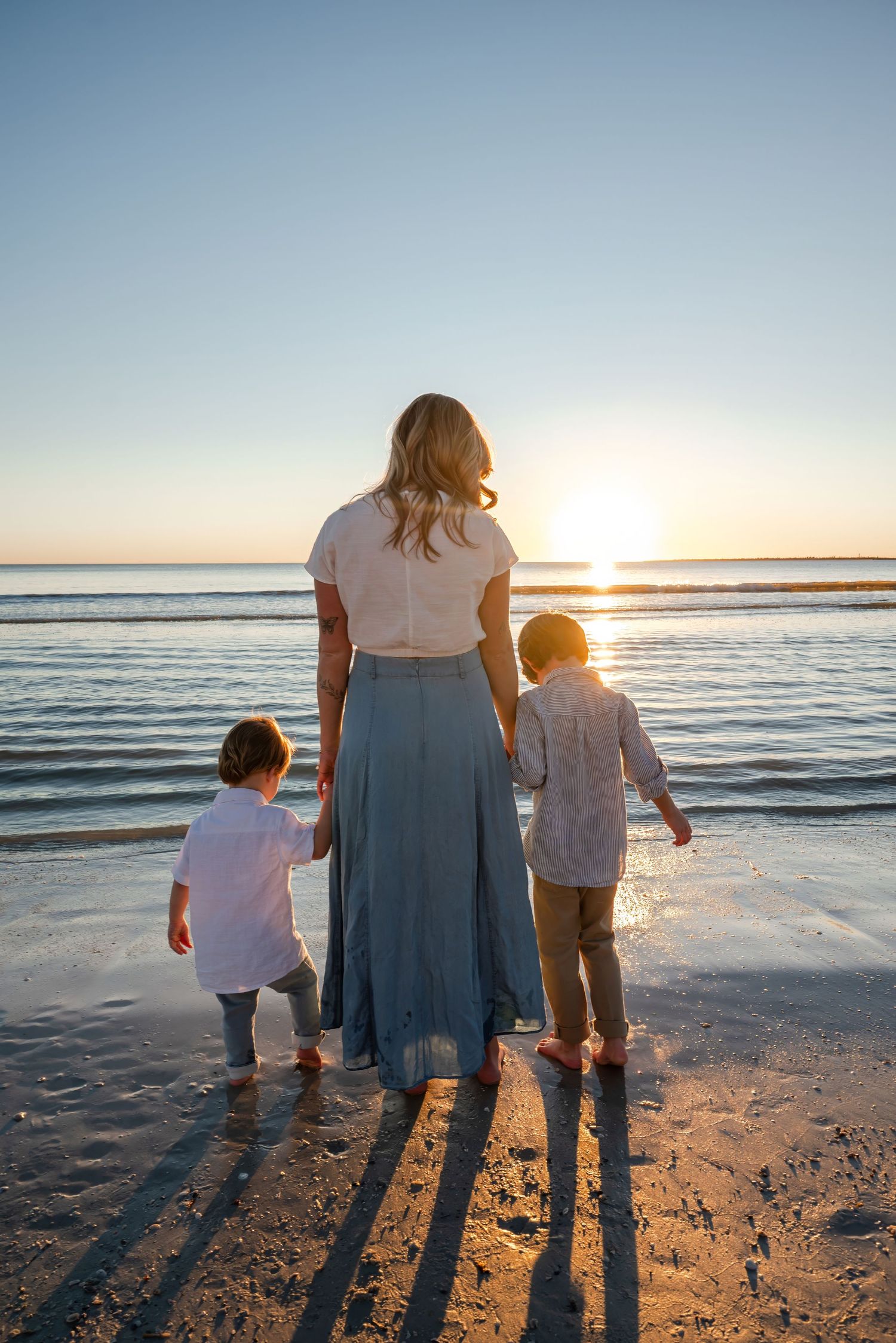 Mother holding hands with two young children facing ocean at sunset.