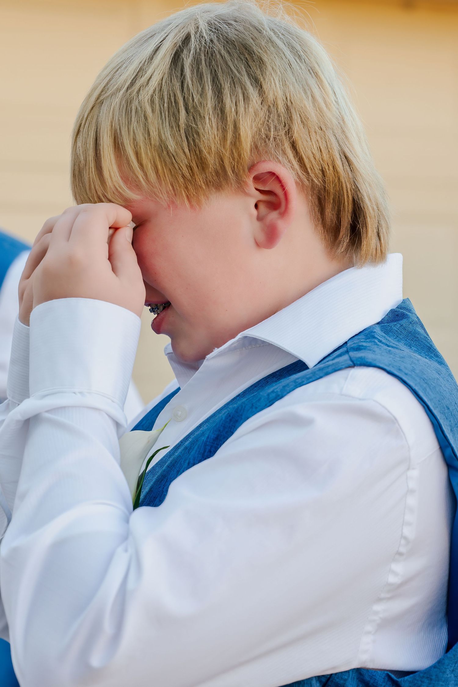 Emotional young blonde boy in a white shirt and blue vest wiping tears from his eyes.