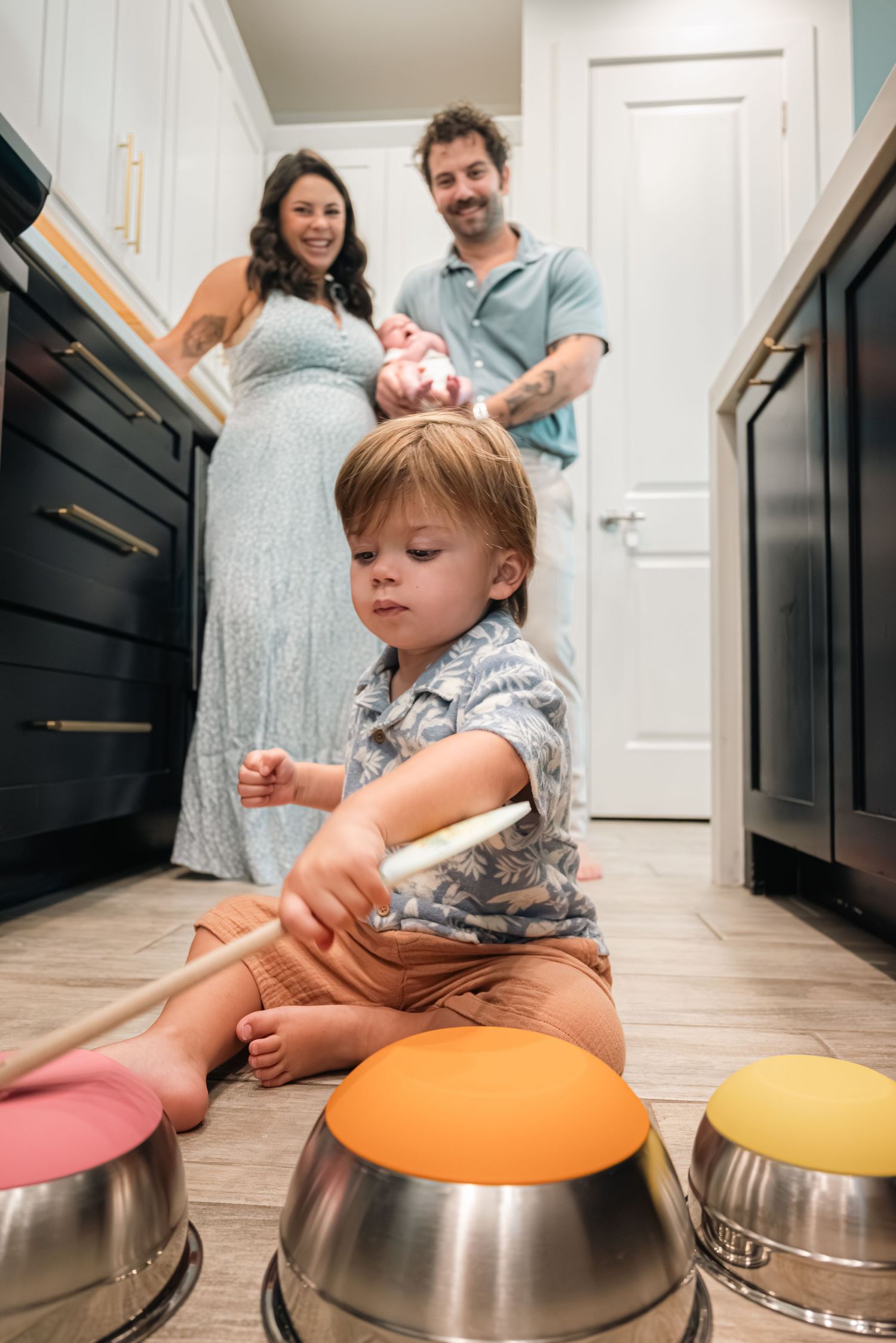 Toddler boy playing with metal bowls and drumsticks on kitchen floor while parents hold newborn baby.